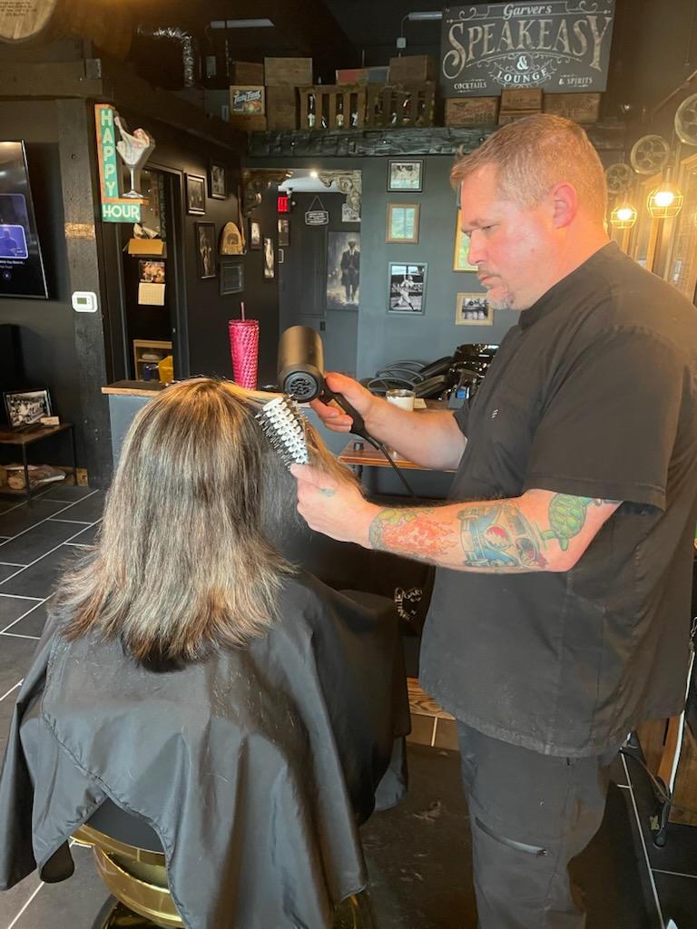A hairstylist with tattoos on his arm is styling a woman's hair in a salon. The woman has shoulder-length hair and a hair accessory.