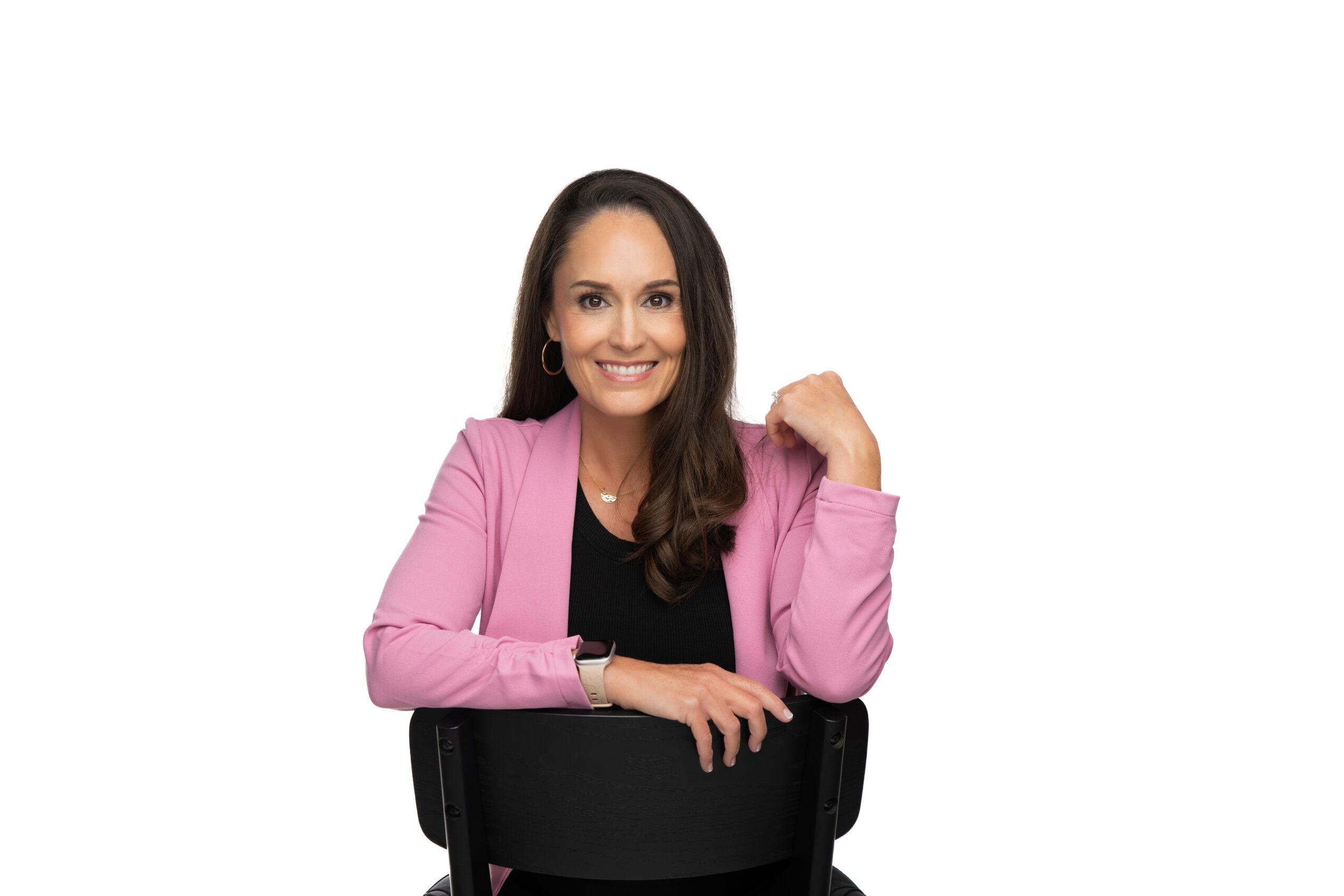 Smiling woman with long dark hair, wearing a pink blazer and black top, sitting on a chair with a white background.