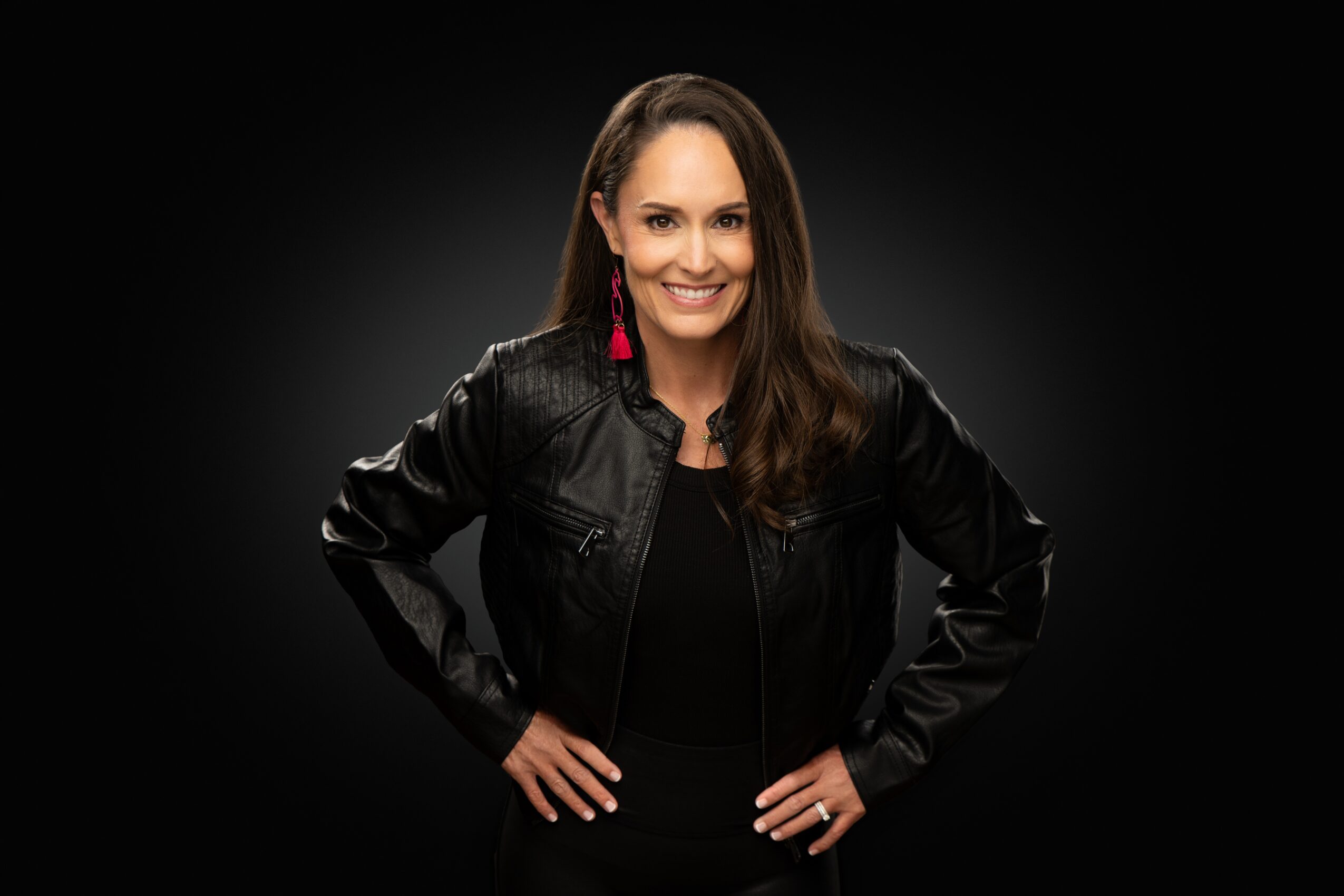Woman with long brown hair smiling, wearing a black leather jacket and pink earrings, standing with hands on hips against a dark background.