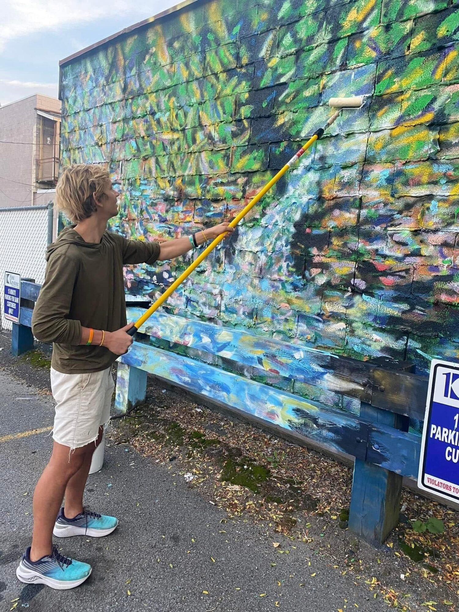 Person painting a colorful mural on a large outdoor wall with a long yellow brush.