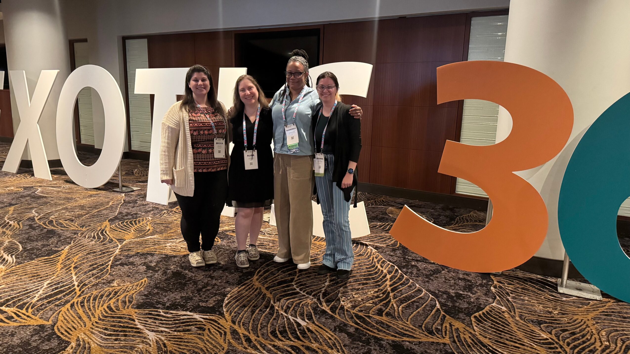Four people standing together in front of large conference signs, smiling, in a carpeted room with a wooden wall.