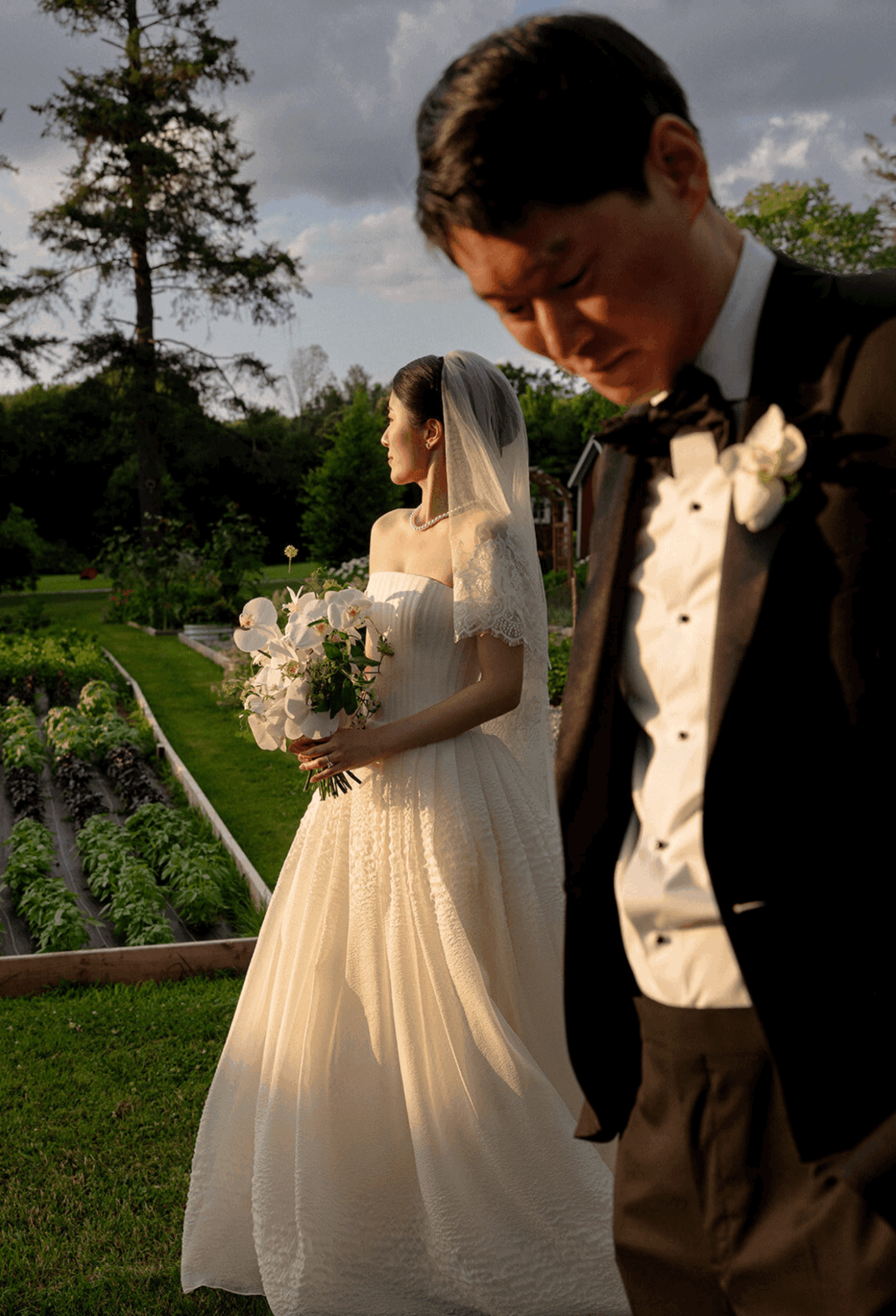Bride in wedding dress holding bouquet, groom in tuxedo with bow tie, outdoors on a sunny day.