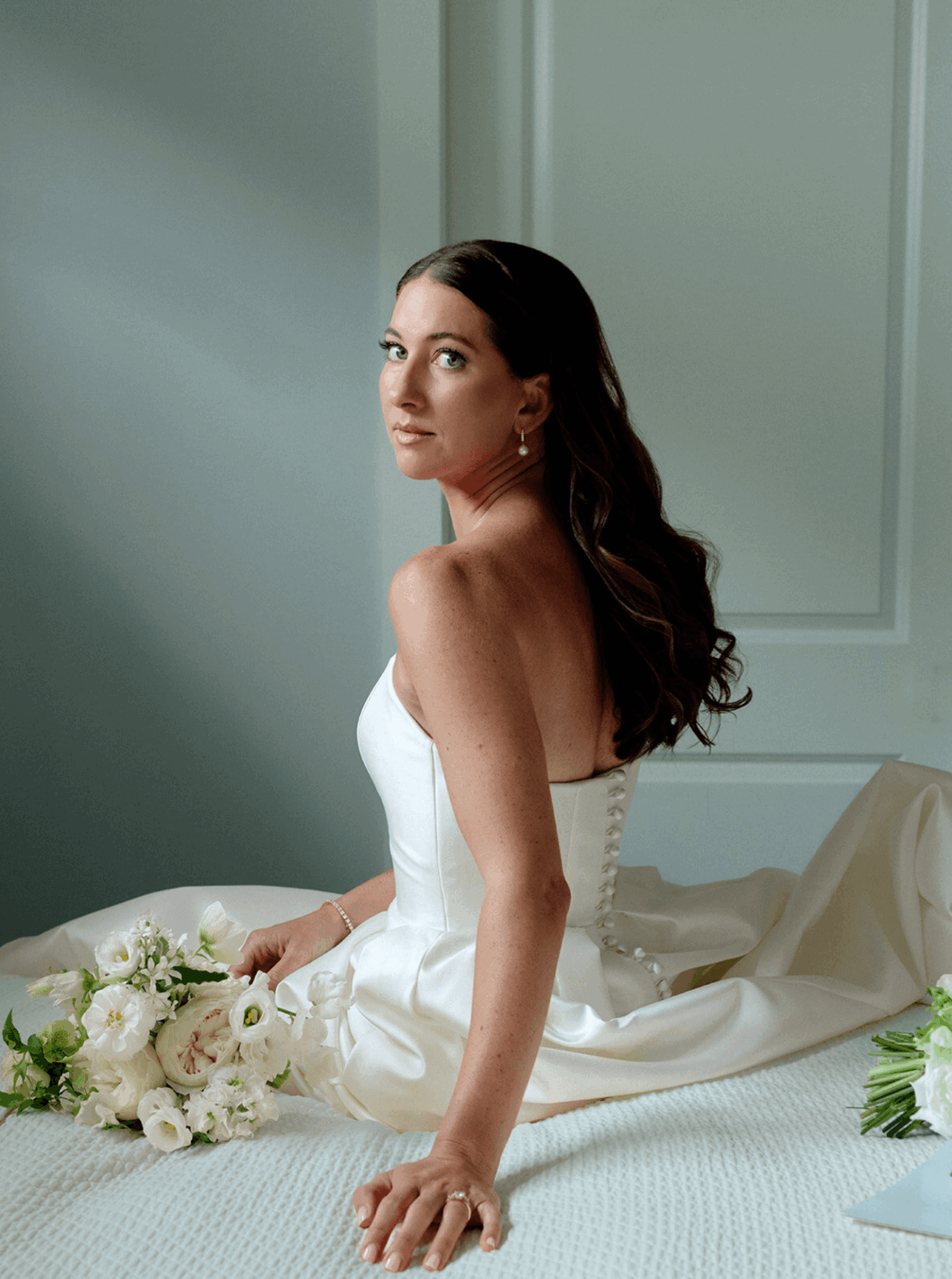 Woman in white dress sitting on bed holding a bouquet of white flowers, looking to the side, in a softly lit room.