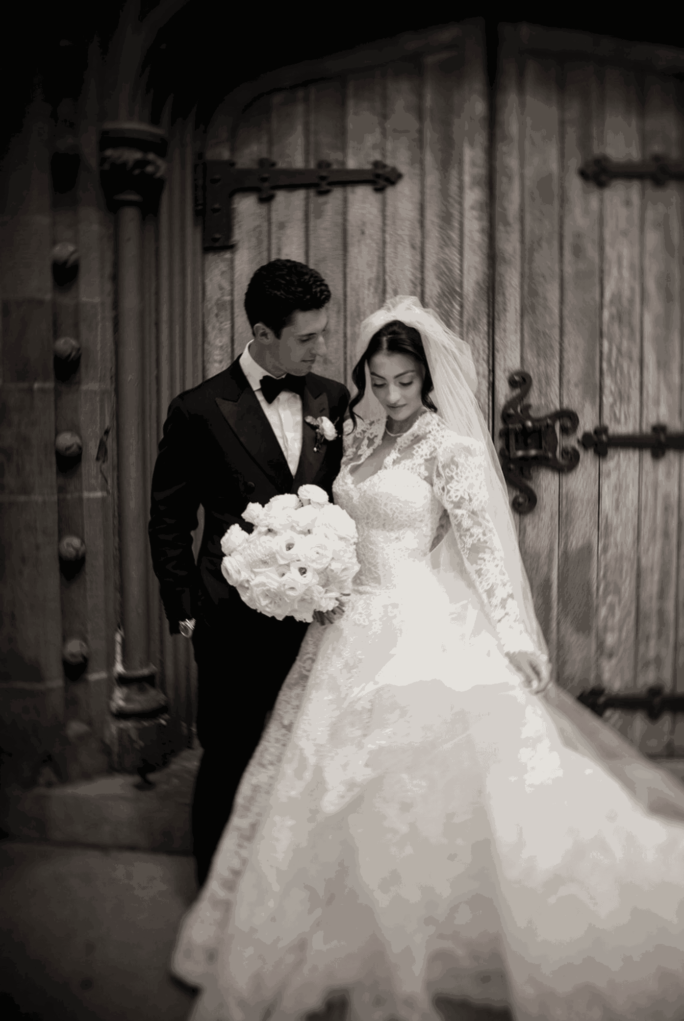Bride and groom in wedding attire standing close, holding a bouquet, in front of large wooden doors, black and white photo.