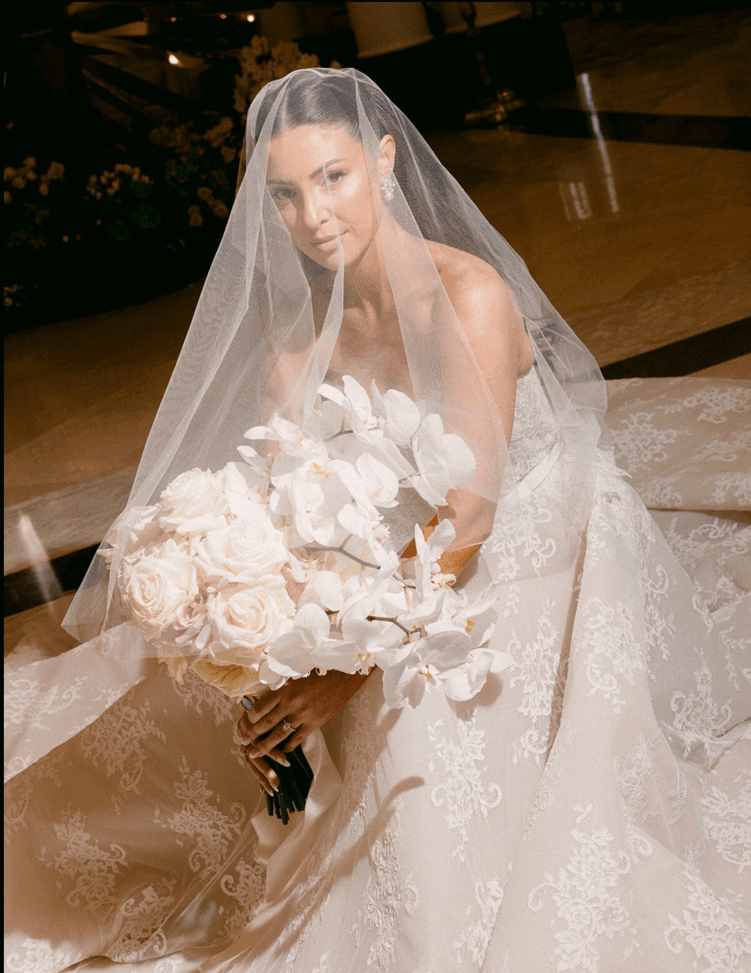 Bride in wedding dress with veil holding a large bouquet of white flowers, sitting on the floor indoors.