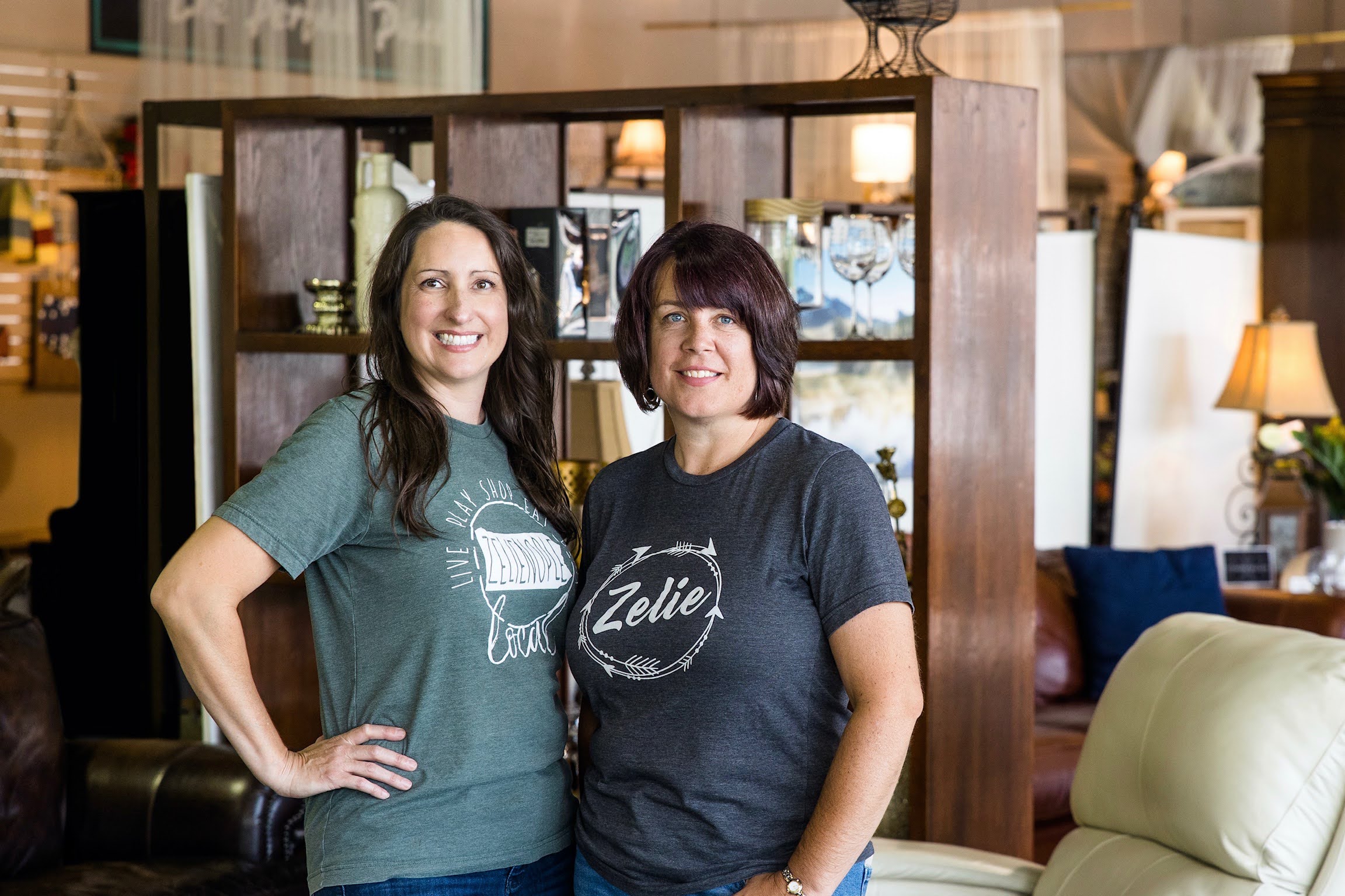 Two women stand side by side in a furniture store, smiling, with shelves and lamps in the background.