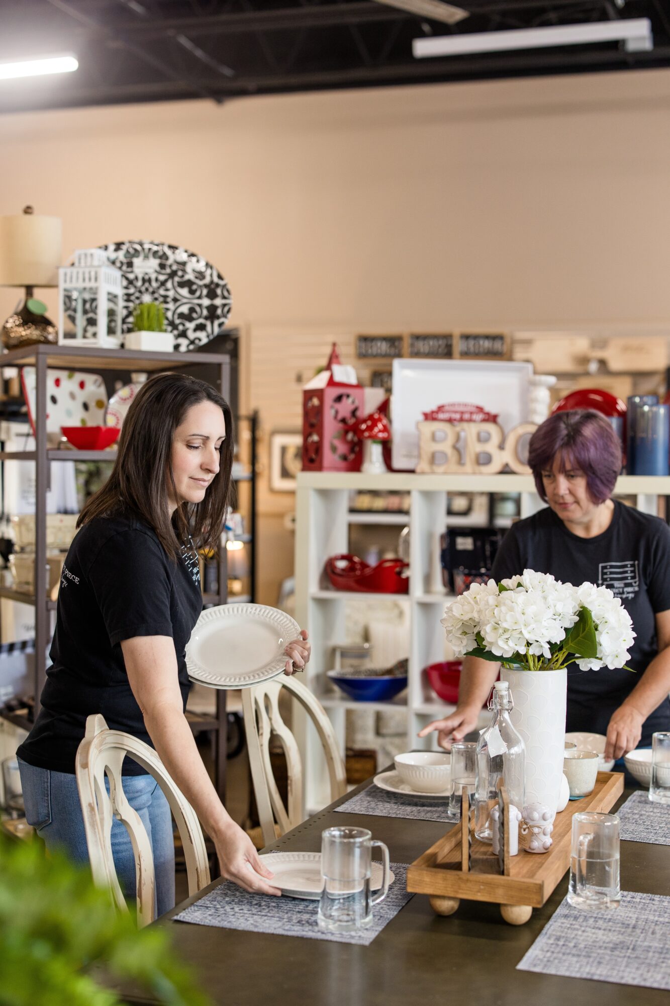 Two women arranging items on a dining table in a store, with shelves of home decor in the background.
