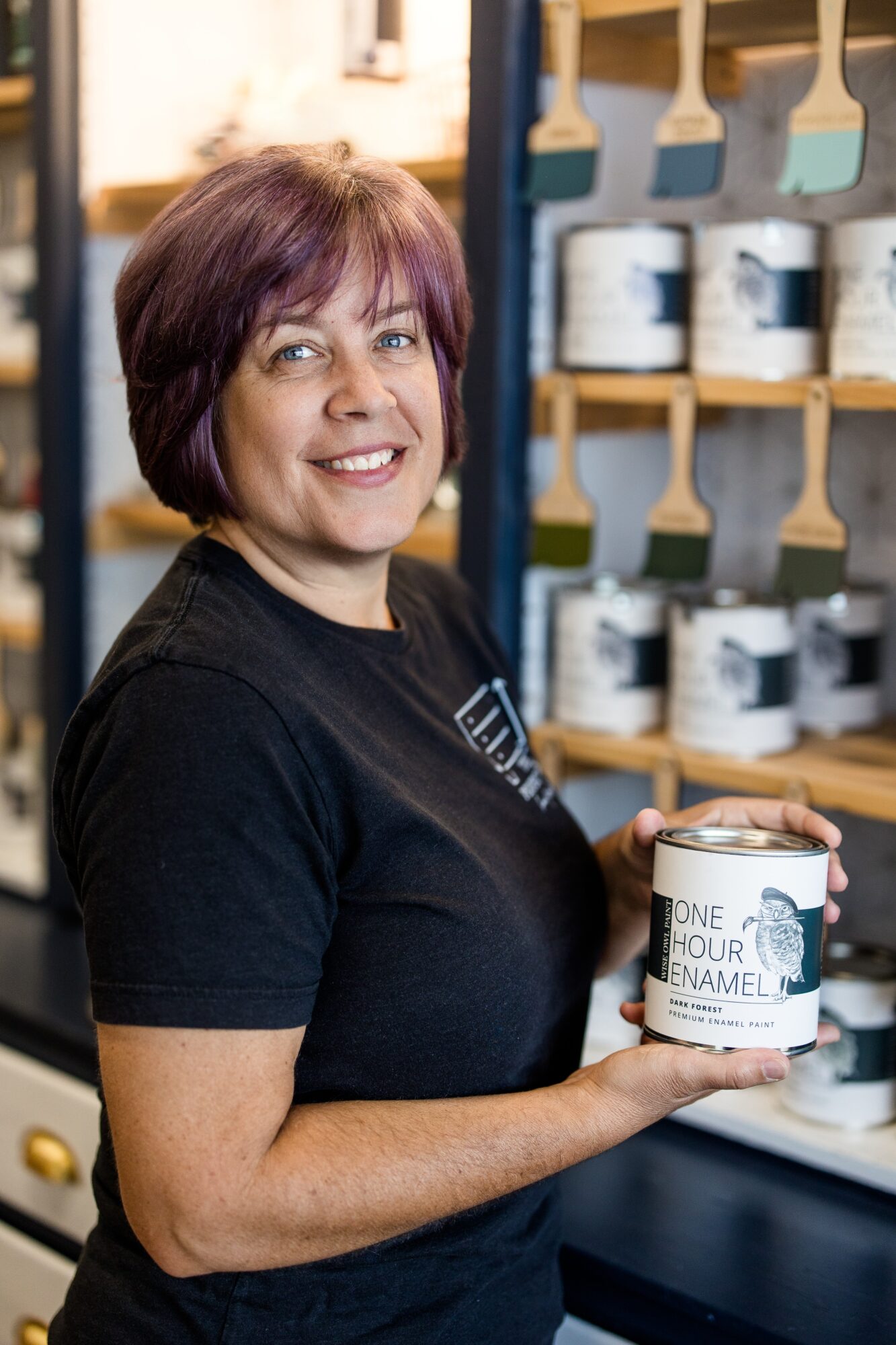 Woman with short purple hair holding a can of paint in a store, shelves with paint cans behind her.