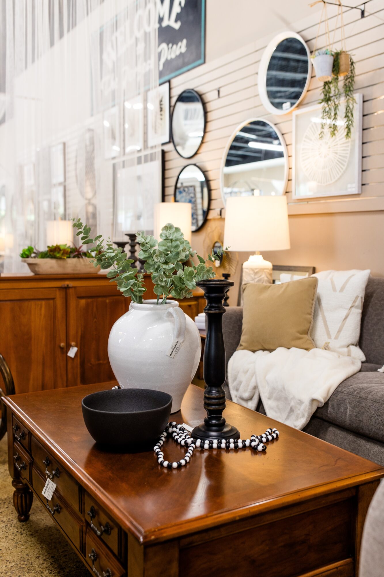 Living room with a wooden table, black bowl, candle holder, and beaded necklace, sofa with pillows, and wall mirrors.