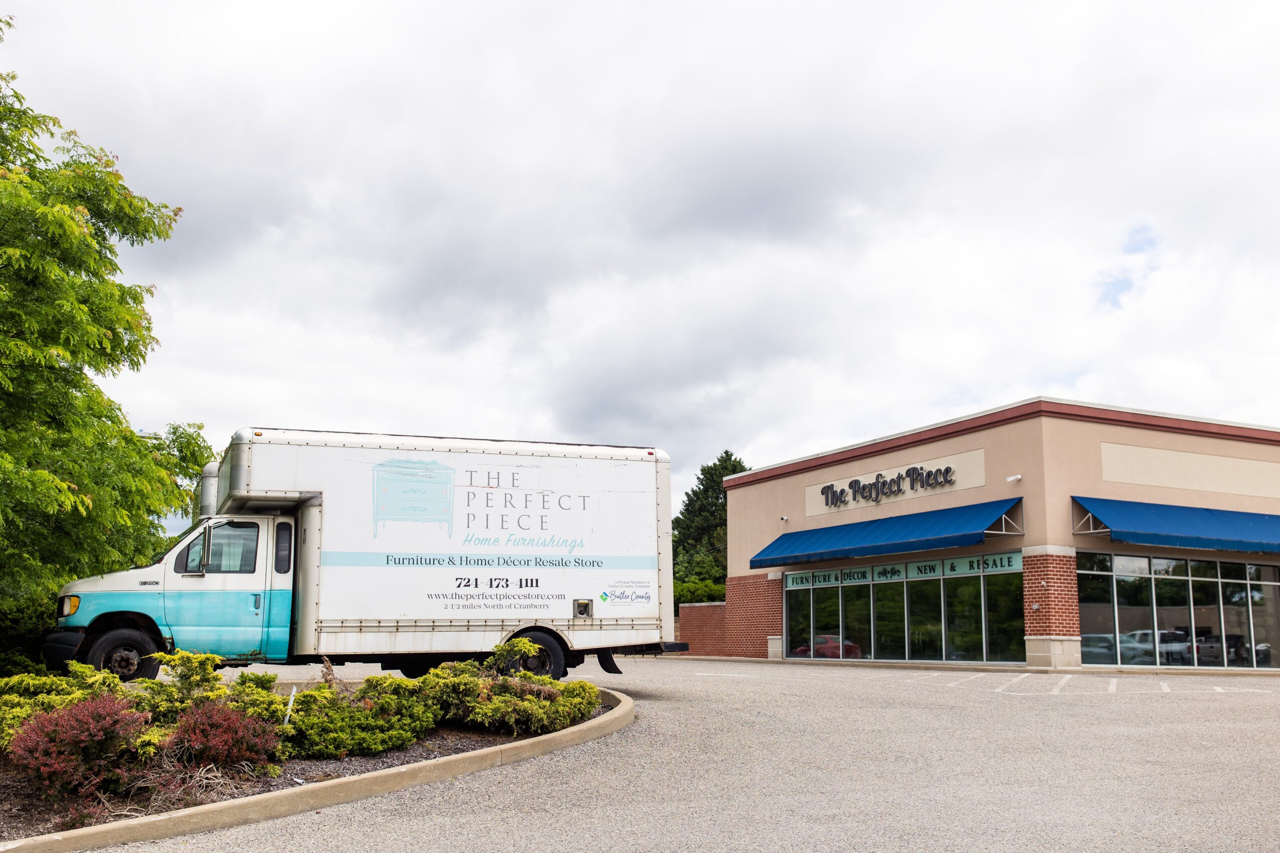 Truck parked outside a retail store with a blue awning, surrounded by greenery and cloudy sky.