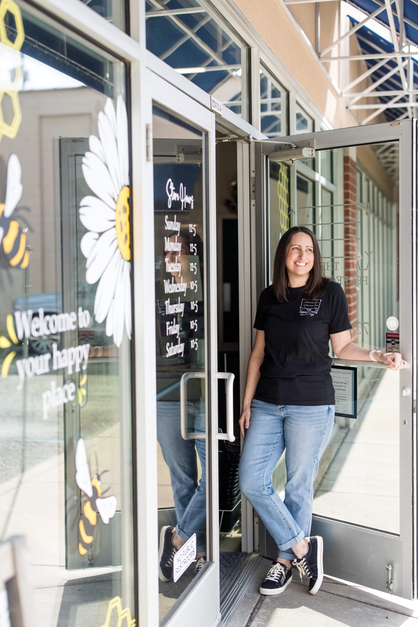 Young woman with glasses and black shirt standing in doorway, smiling, with glass and floral decorations outside.
