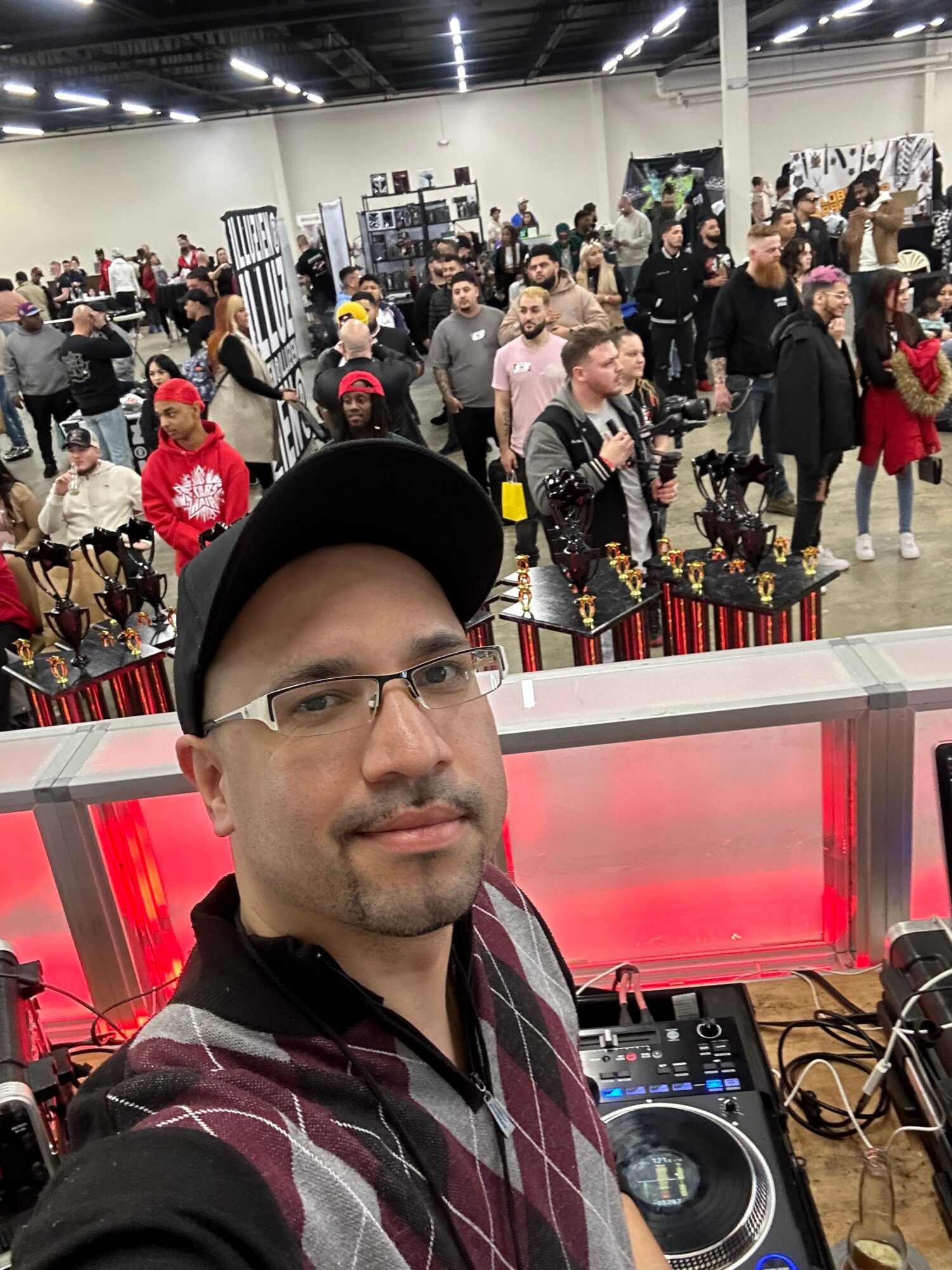Person wearing glasses, a cap, and a plaid shirt taking a selfie at a crowded indoor event with people in the background.