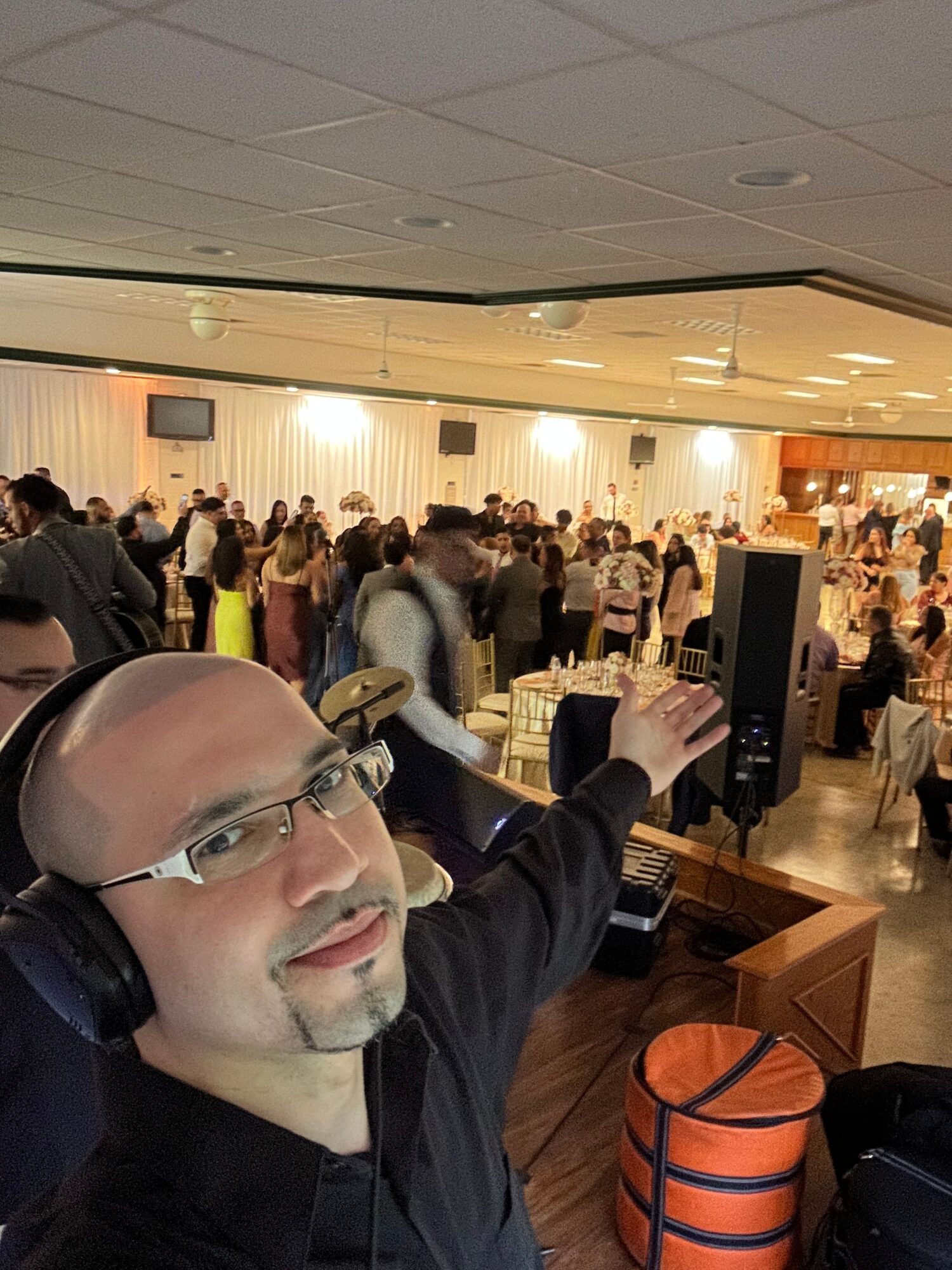 Bald person with glasses and headphones smiling at a social event, with a crowd in the background.