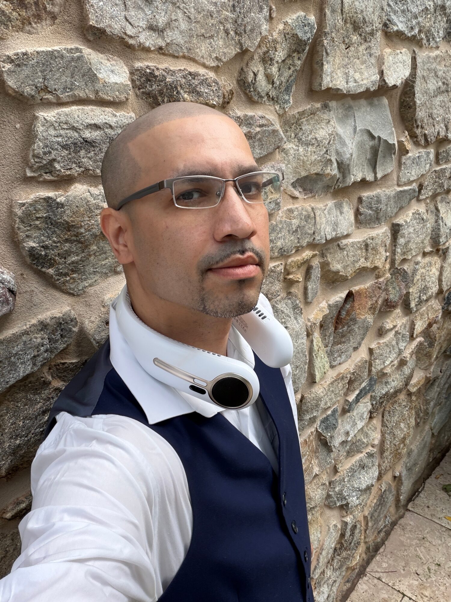 Man with glasses and headphones around neck standing against a stone wall, looking to the side.