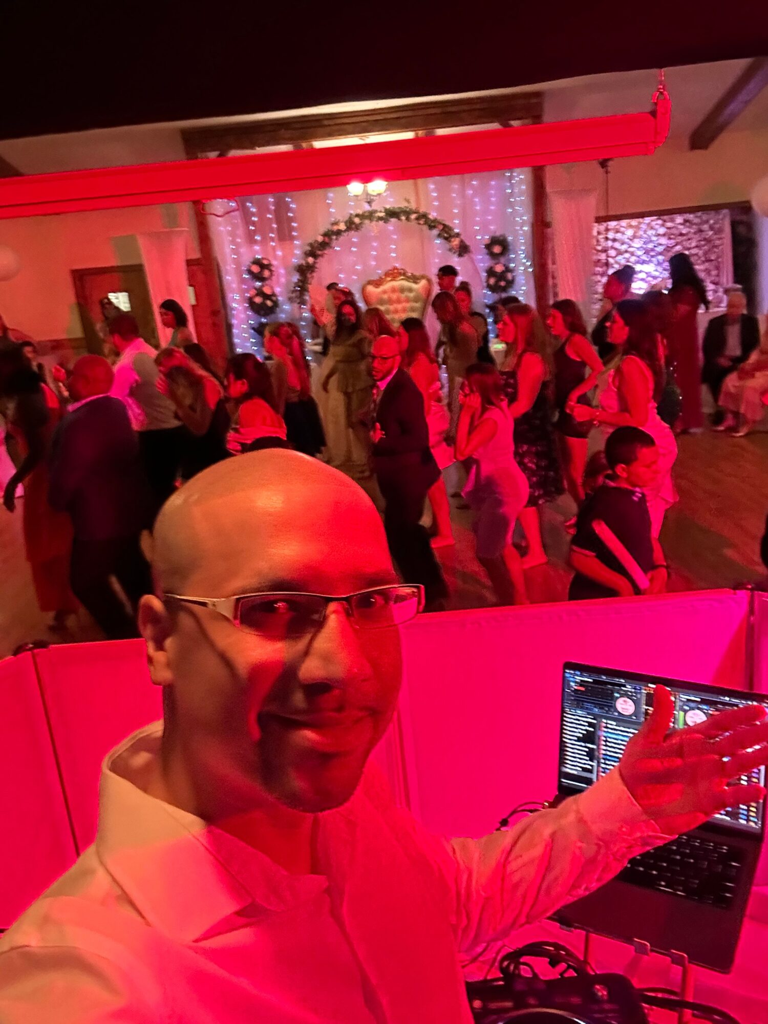 Person with glasses and bald head in a white shirt, taking a selfie at a party with people in the background.