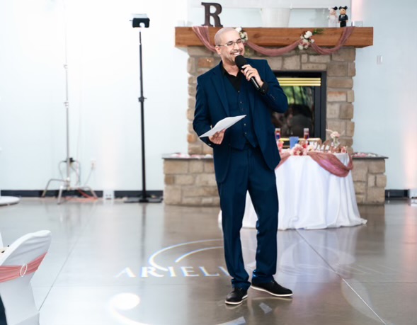 Man in suit holding microphone and paper, standing in front of fireplace with decorations, speaking to audience.