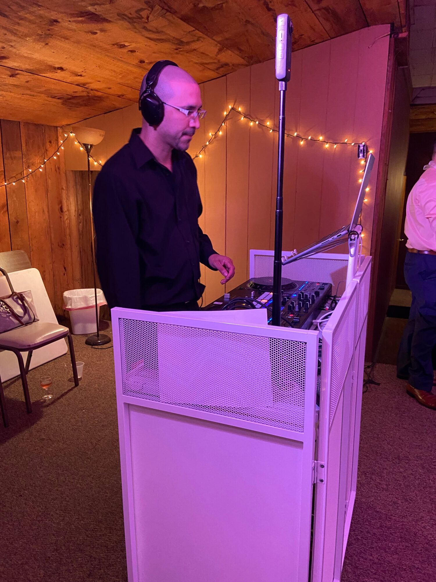 Person wearing headphones stands behind a white DJ booth with a microphone, in a room with wooden walls and string lights.