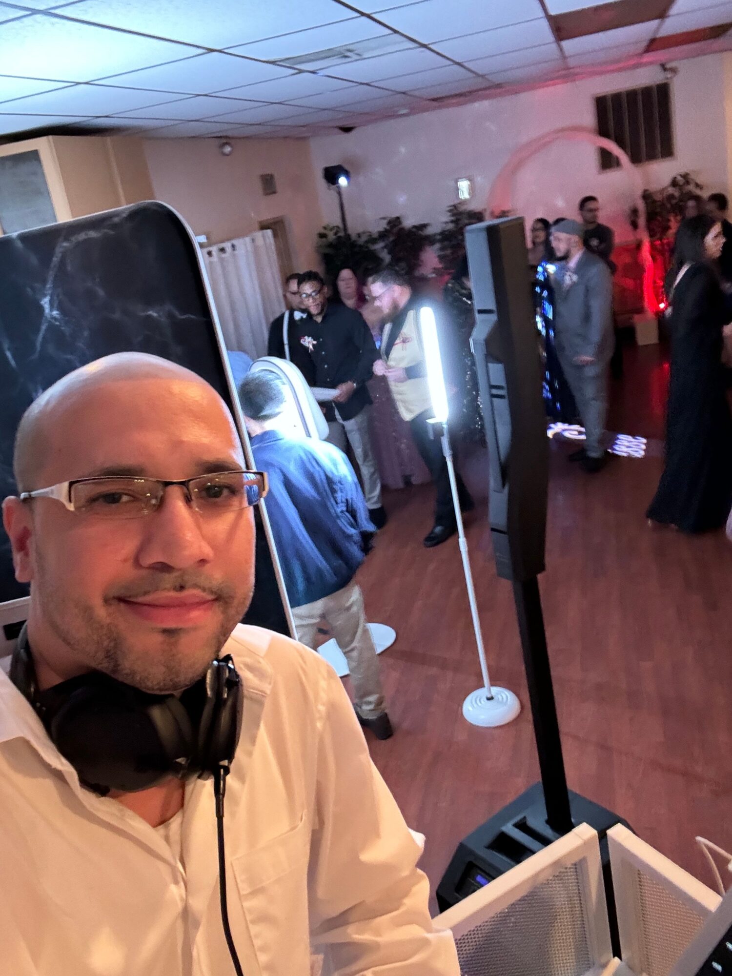 Man with glasses and headphones in a room with people and equipment, facing camera, smiling.