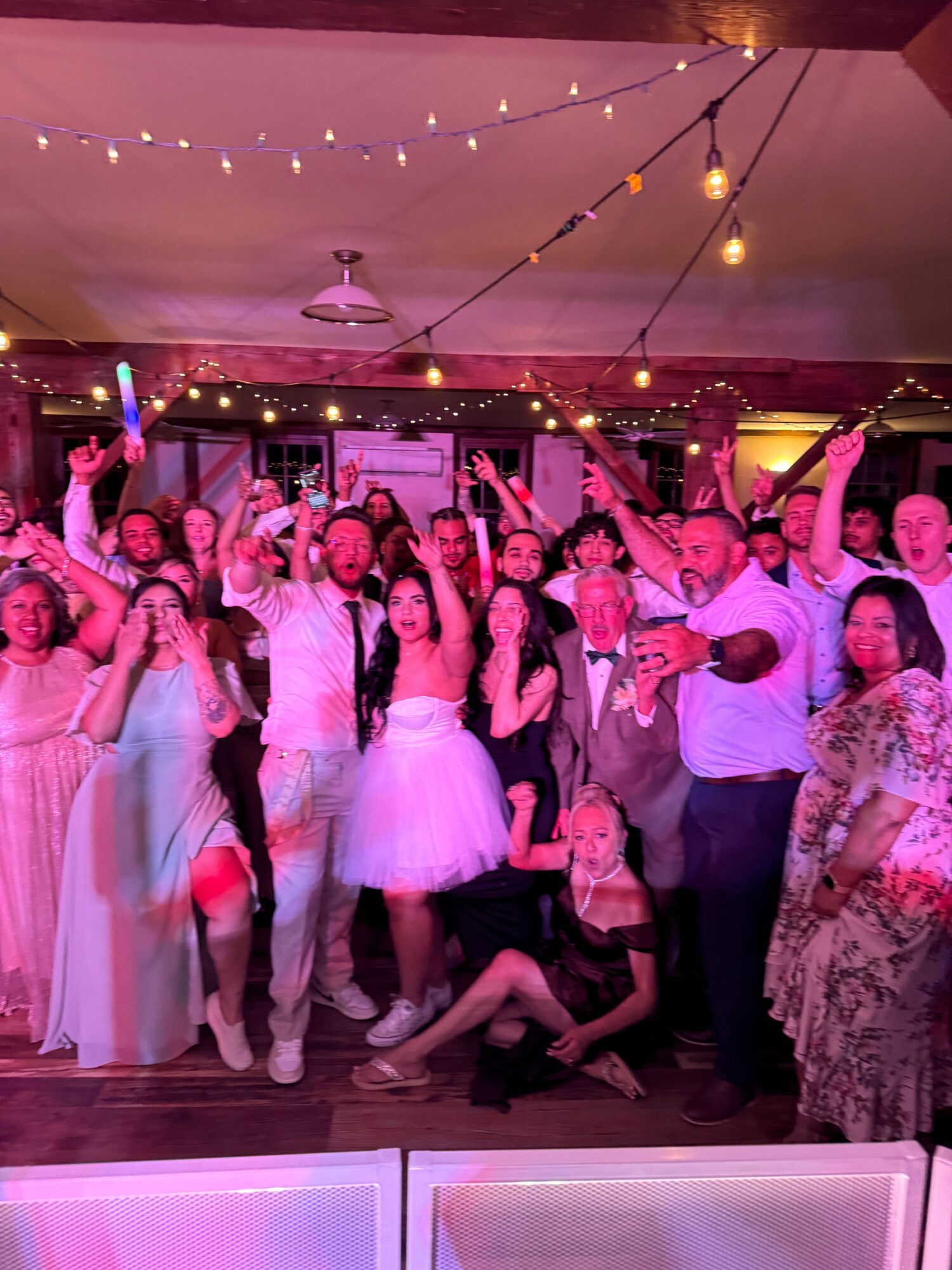 Group of people at a celebration or party, some holding drinks, with pink and yellow lighting, and string lights overhead.