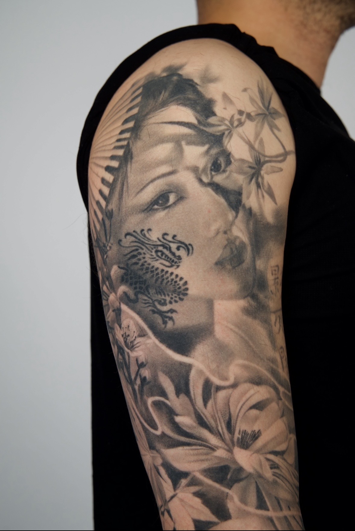 Tattoo of a woman's face with floral and dragon elements on upper arm, black and gray shading, detailed design.