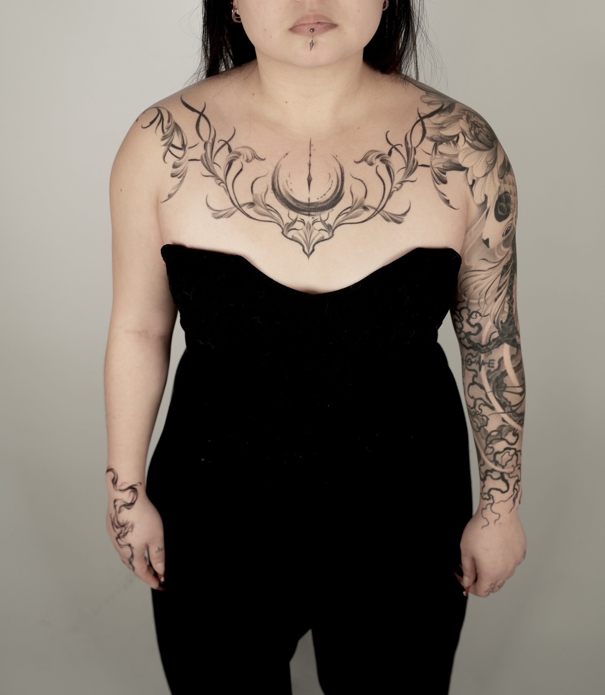Person with tattoos on chest, arms, and hands wearing a black strapless outfit, facing forward, against a plain background.