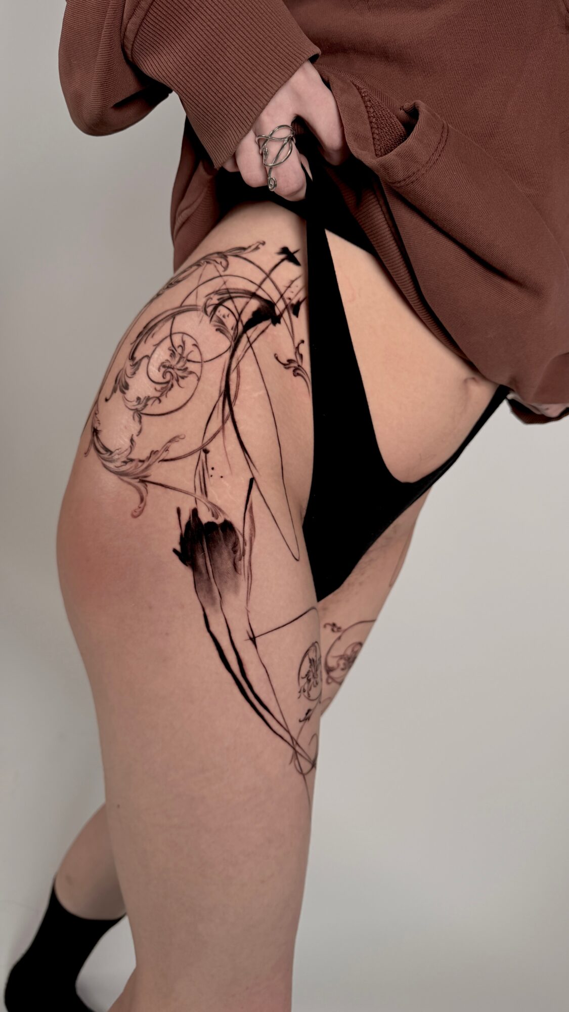 Person with a large, abstract black tattoo on thigh, wearing black underwear and a brown top, standing against a plain background.