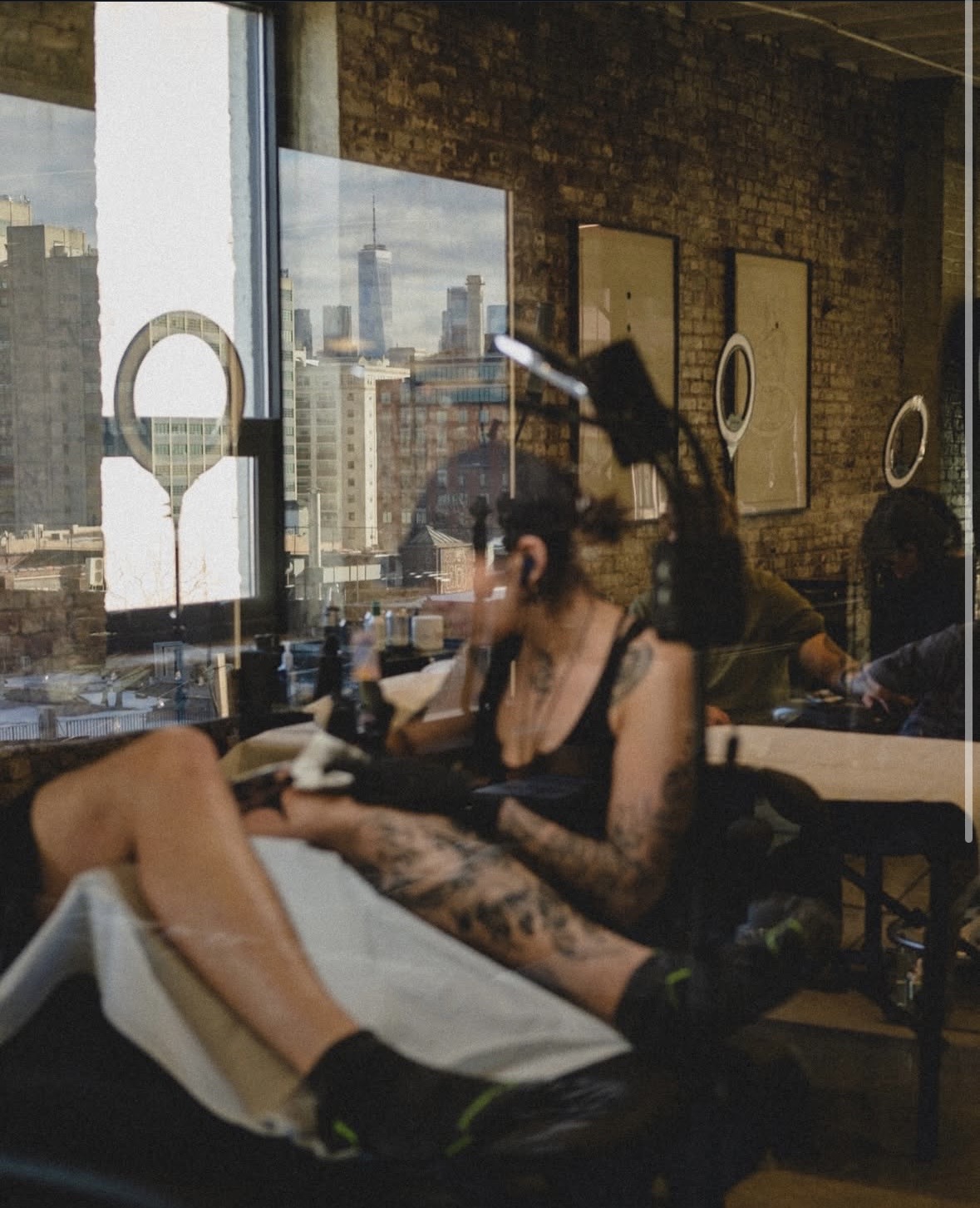 Tattooed person receiving a manicure in a salon with large windows and cityscape view.