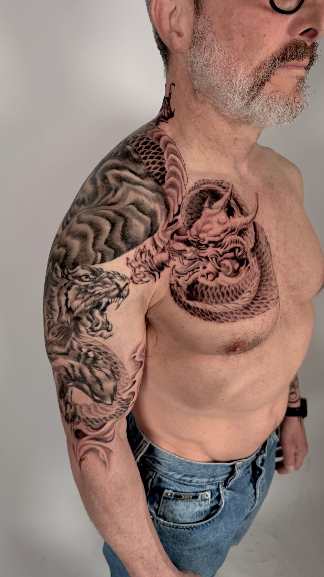 Man with a large tattoo of a dragon and a tiger on his shoulder and chest, wearing jeans, shirtless.