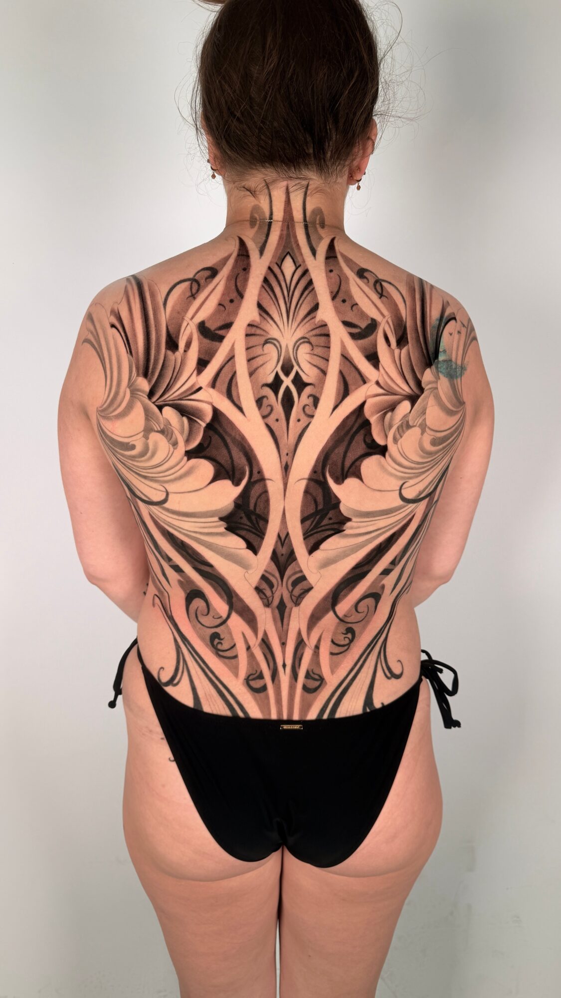 Back view of a person with a large symmetrical tattoo covering the shoulders and back, wearing black bottoms.
