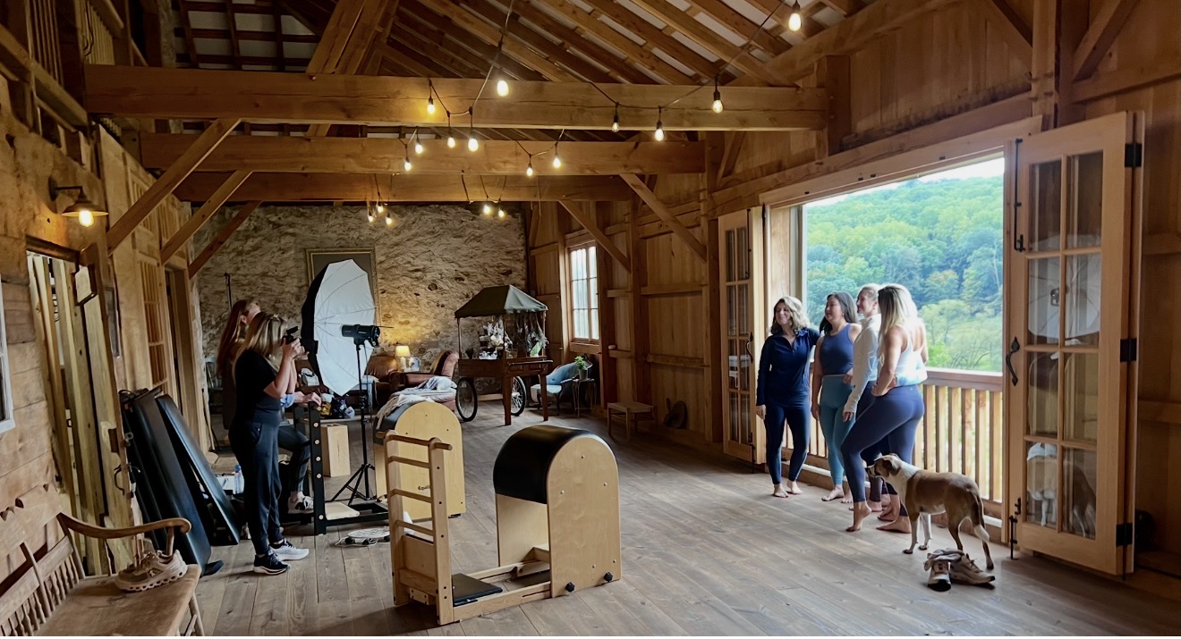 People inside a wooden barn with large open doors, some with dogs, and a scenic view outside, warm lighting, rustic interior.