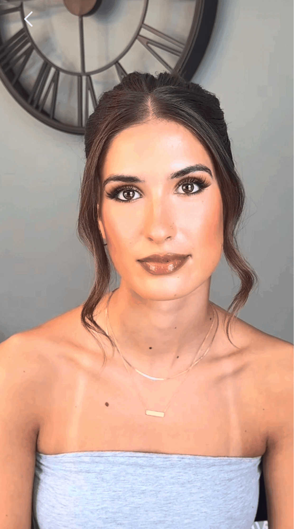 Young woman with dark hair styled in loose waves, wearing makeup and a strapless top, standing in front of a clock.