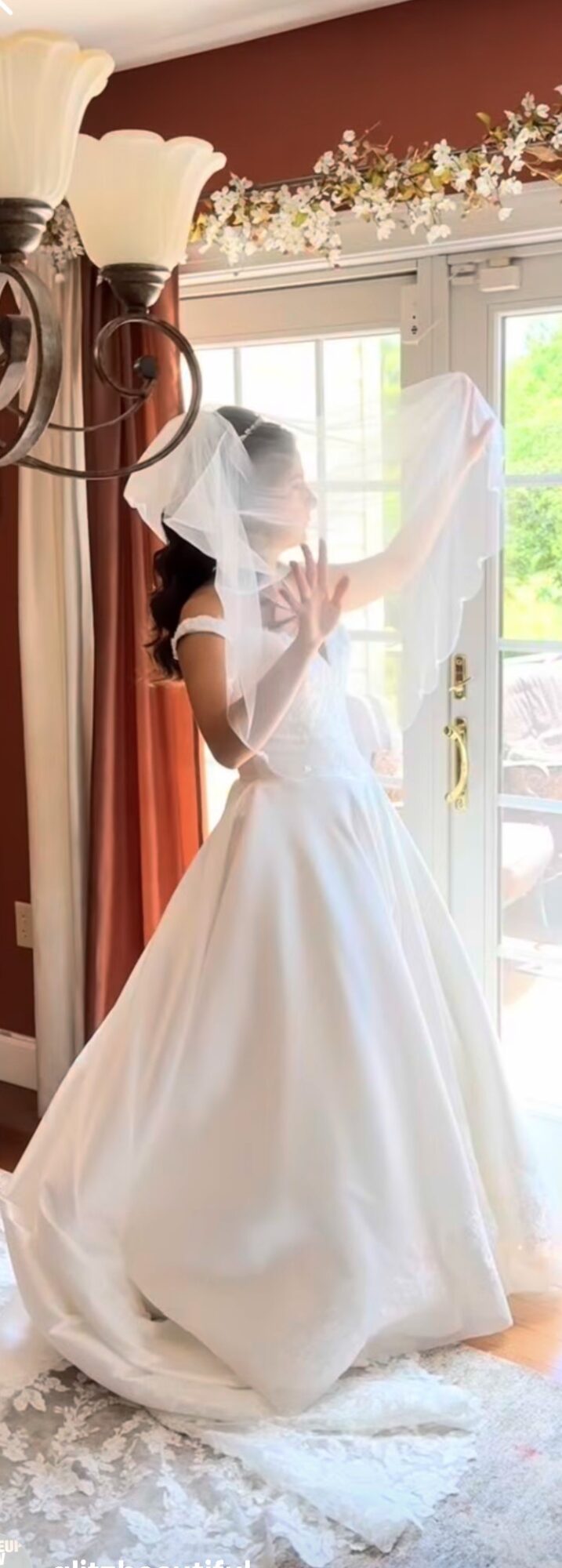 A bride in a white wedding dress and veil stands near a window, holding a glass door handle, with floral decorations above.