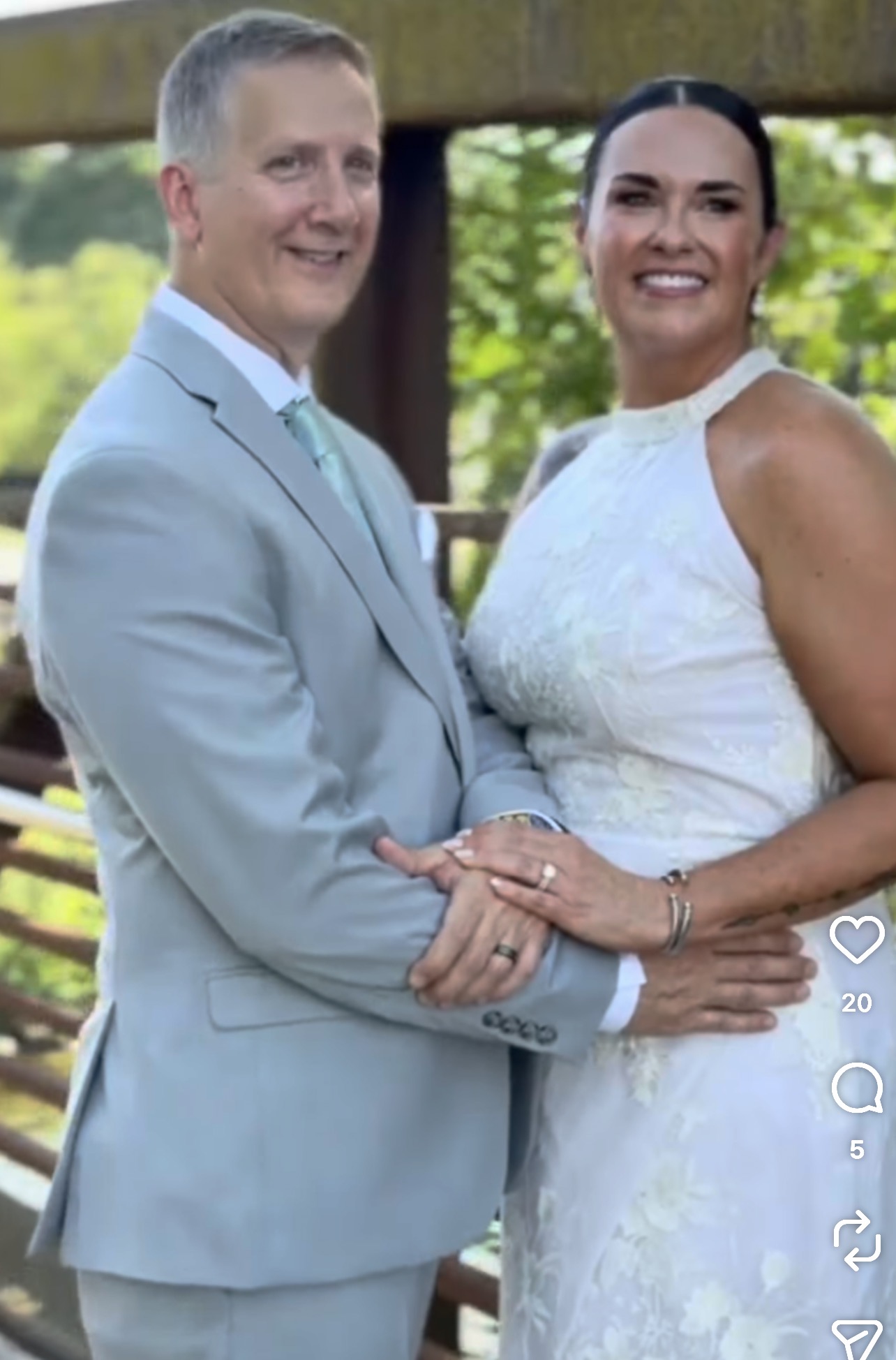 A smiling couple in formal attire, standing outdoors with trees and a wooden structure in the background.