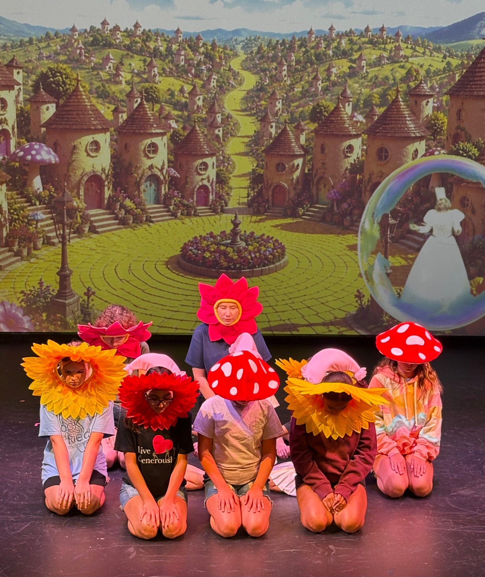 Five children in costumes with large flower and mushroom hats sit on stage, with a castle backdrop and a large glass slipper.