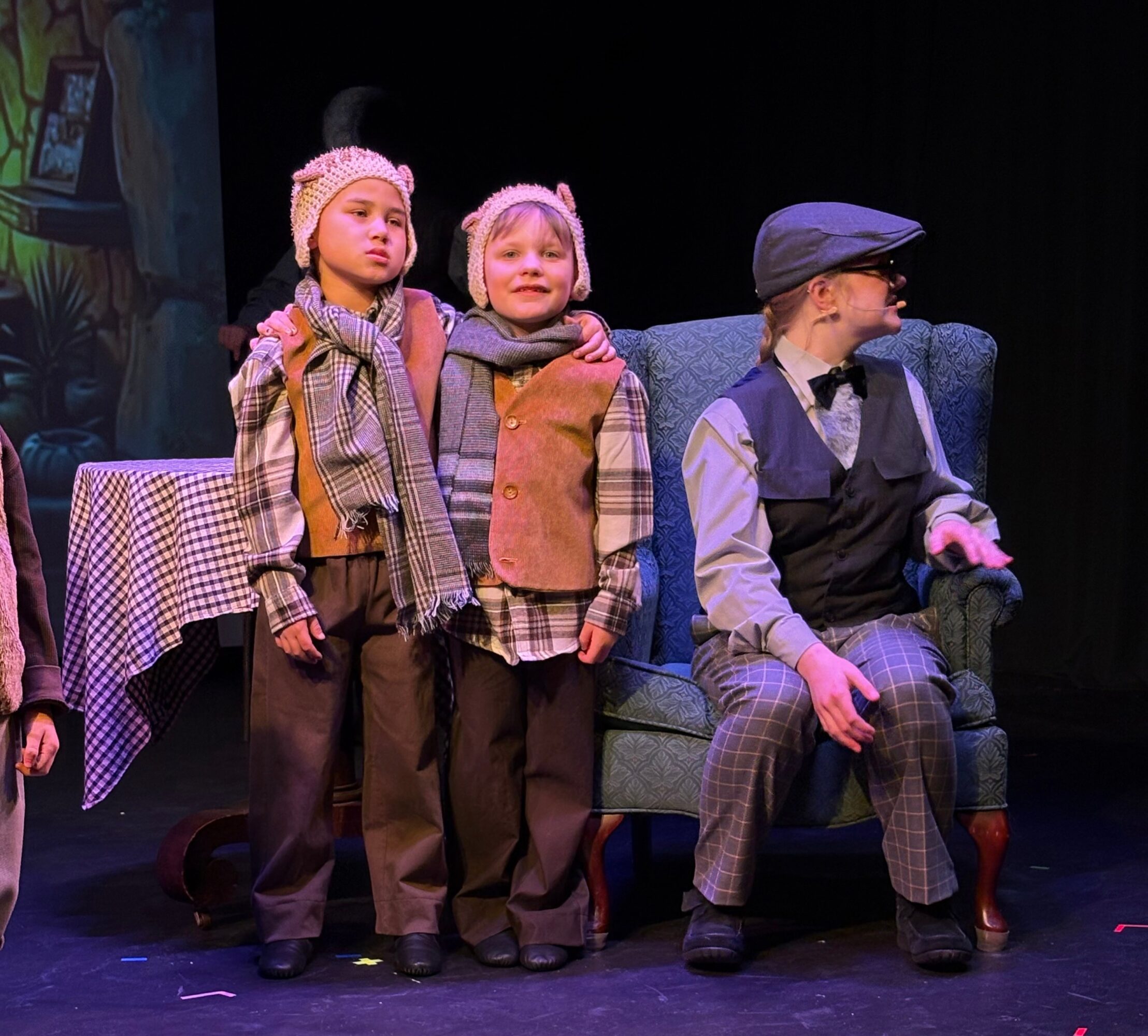 Three children in costumes on stage, two standing and one sitting on a blue armchair, with a table nearby.
