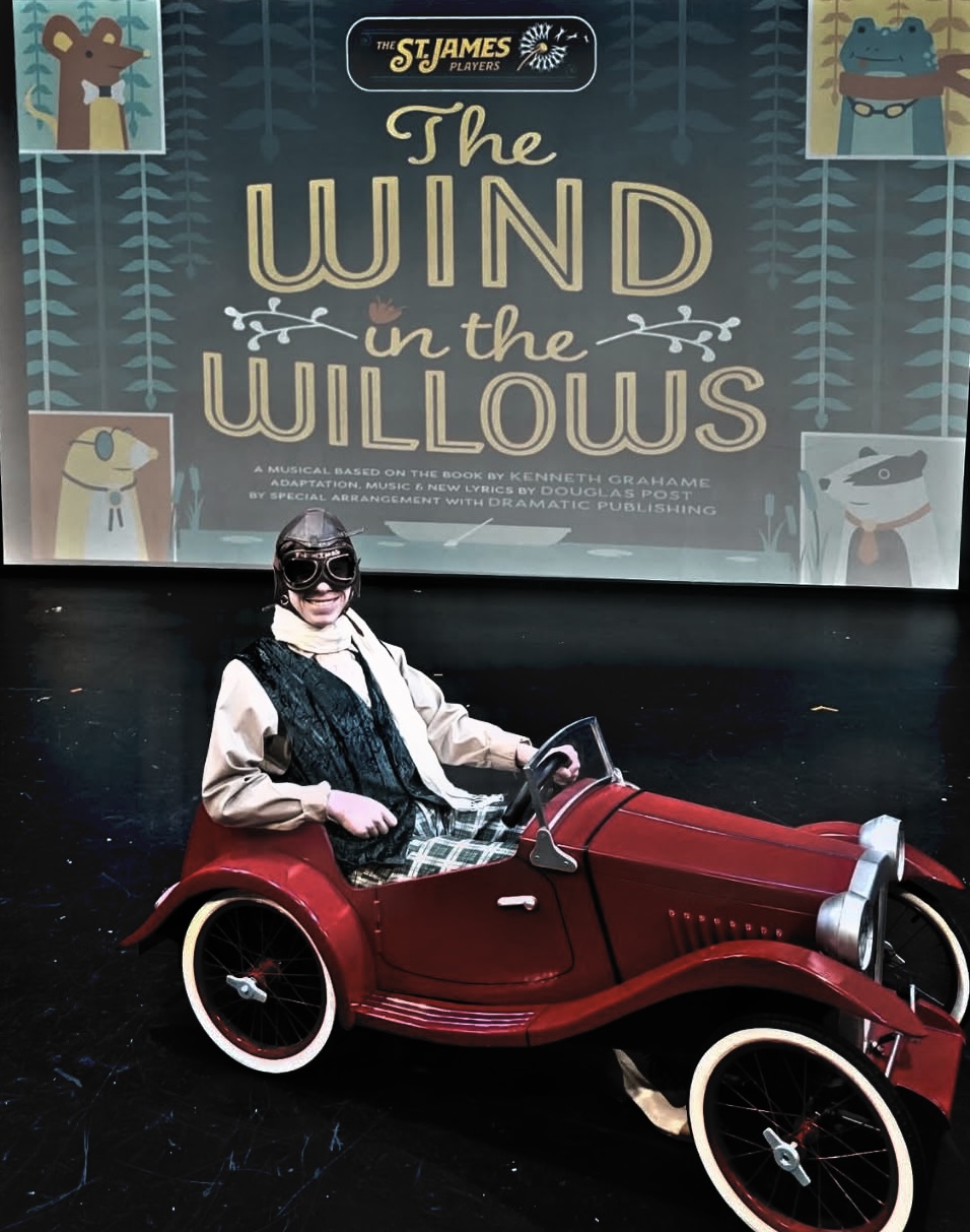 Person sitting in a red vintage-style car on stage with a backdrop reading 'The Wind in the Willows' and illustrations of animals.