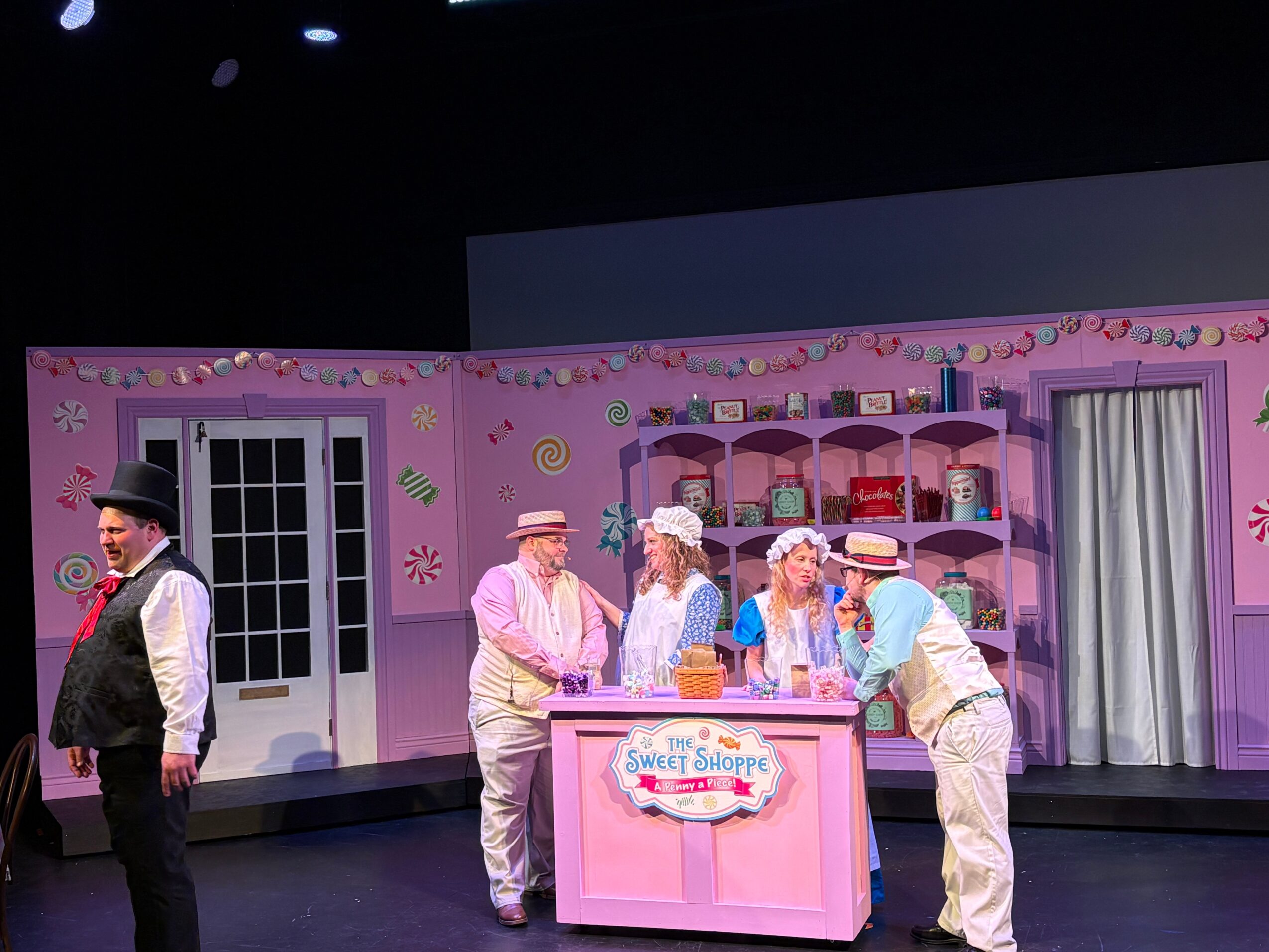 Stage scene with people in costumes around a pink shop counter, set in a colorful room with shelves and a door.