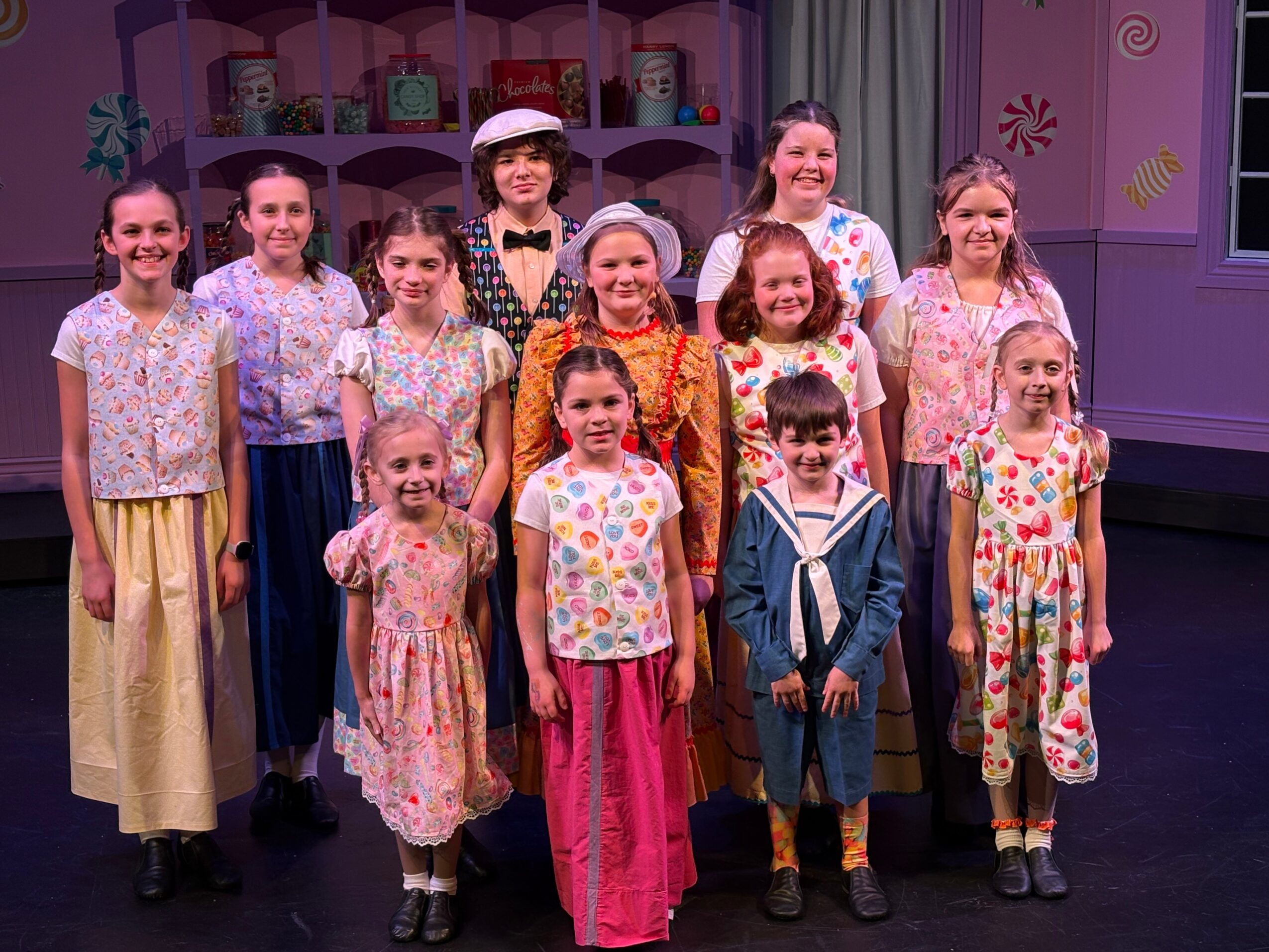 Group of children and an adult on stage, smiling, in colorful costumes, with a set background. 20 words.