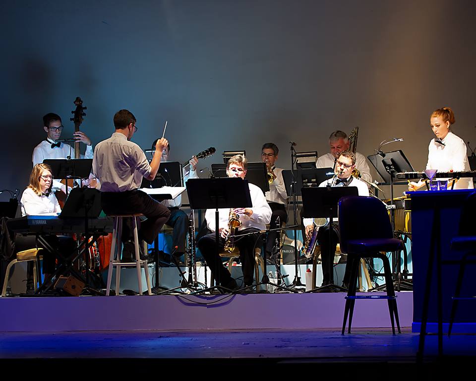 Orchestra performing on stage with conductor, musicians playing instruments, stage lighting, and empty chairs in front.