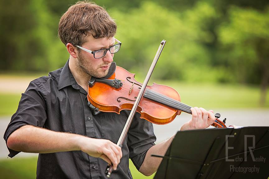 Young man playing violin outdoors, reading sheet music, with green trees in background.