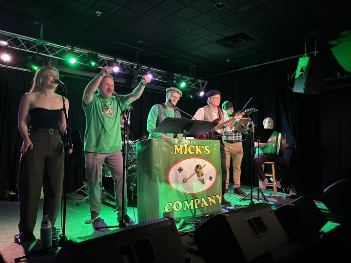 Five musicians perform on stage with green lighting, one singing, others playing instruments, and a banner reading 'MICK'S COMPANY'.