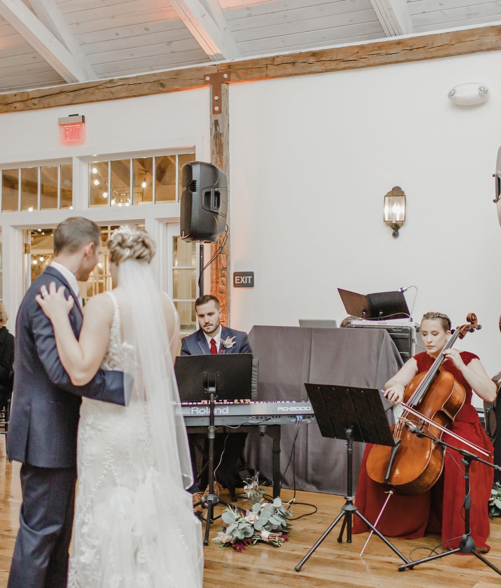 People at a wedding reception with a musician playing piano and a cellist performing indoors, decorated with flowers.