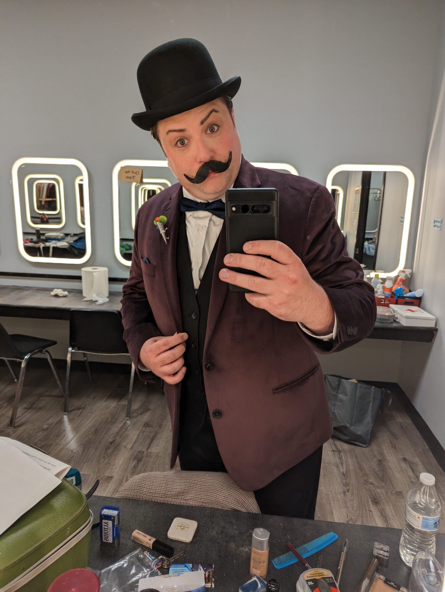 Person dressed as a vintage performer with a top hat, mustache, and tuxedo, taking a selfie in a dressing room.