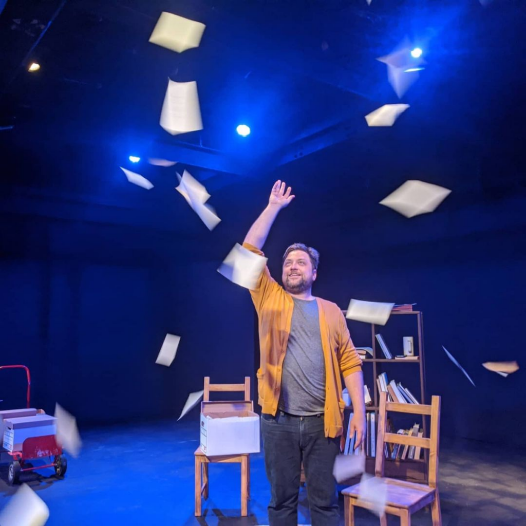 Man standing on stage with books and chairs, papers falling from above, dark background, blue lighting.