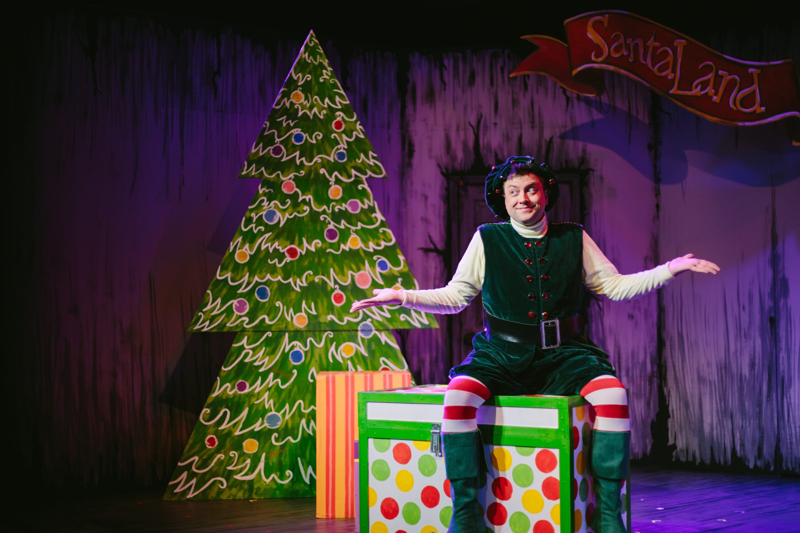 Person dressed as a clown sitting on a colorful box with a decorated Christmas tree and a stage background.