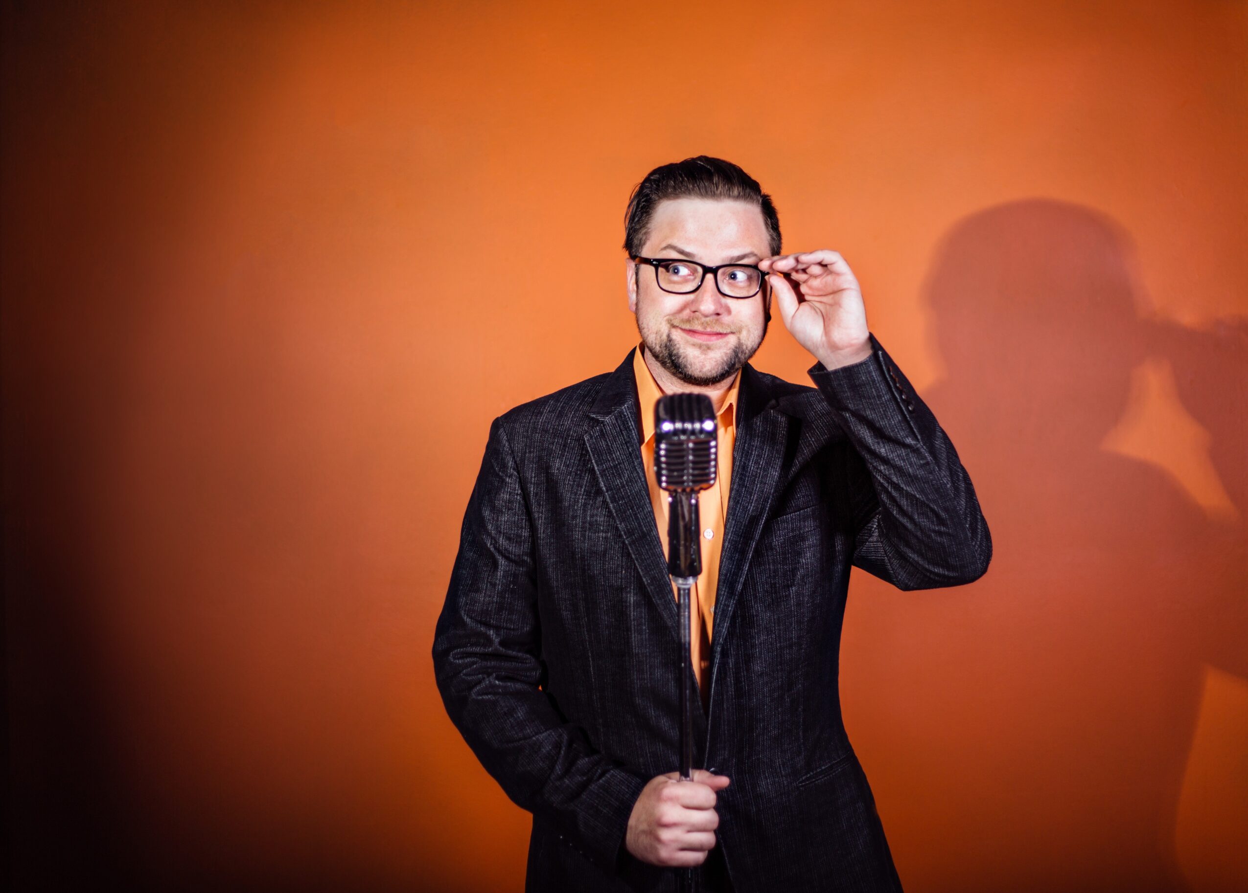 Man with glasses and beard adjusting his glasses, standing in front of an orange background, holding a microphone.