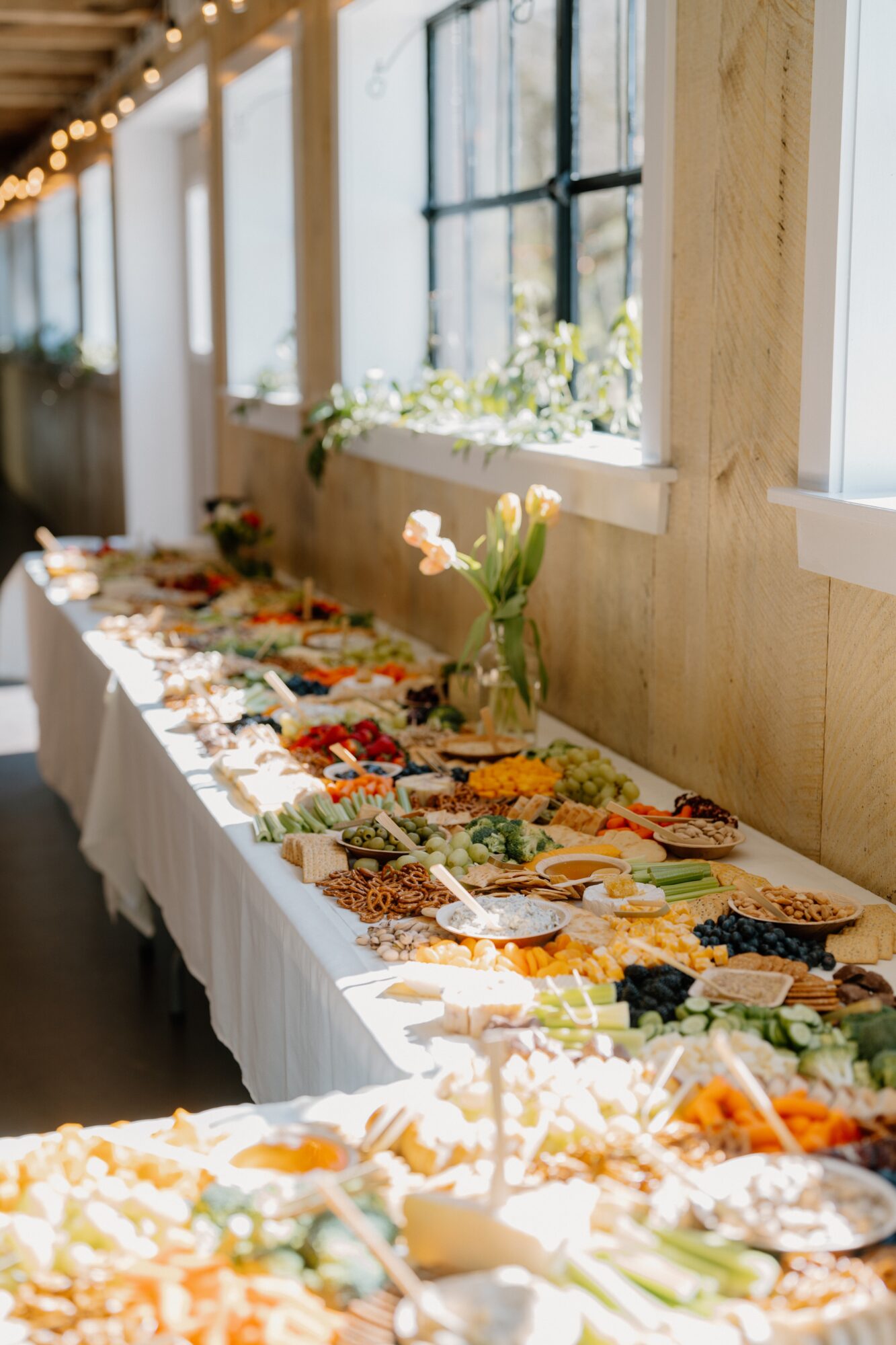 Long table with food and flowers near windows, sunlight streaming in, decorated for an event.