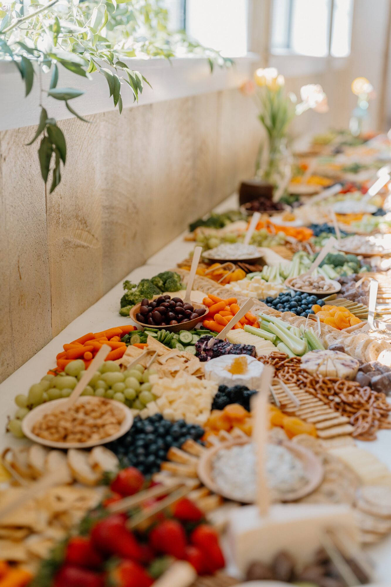 Assorted fruits, desserts, and snacks arranged on a table with a window and greenery in the background.