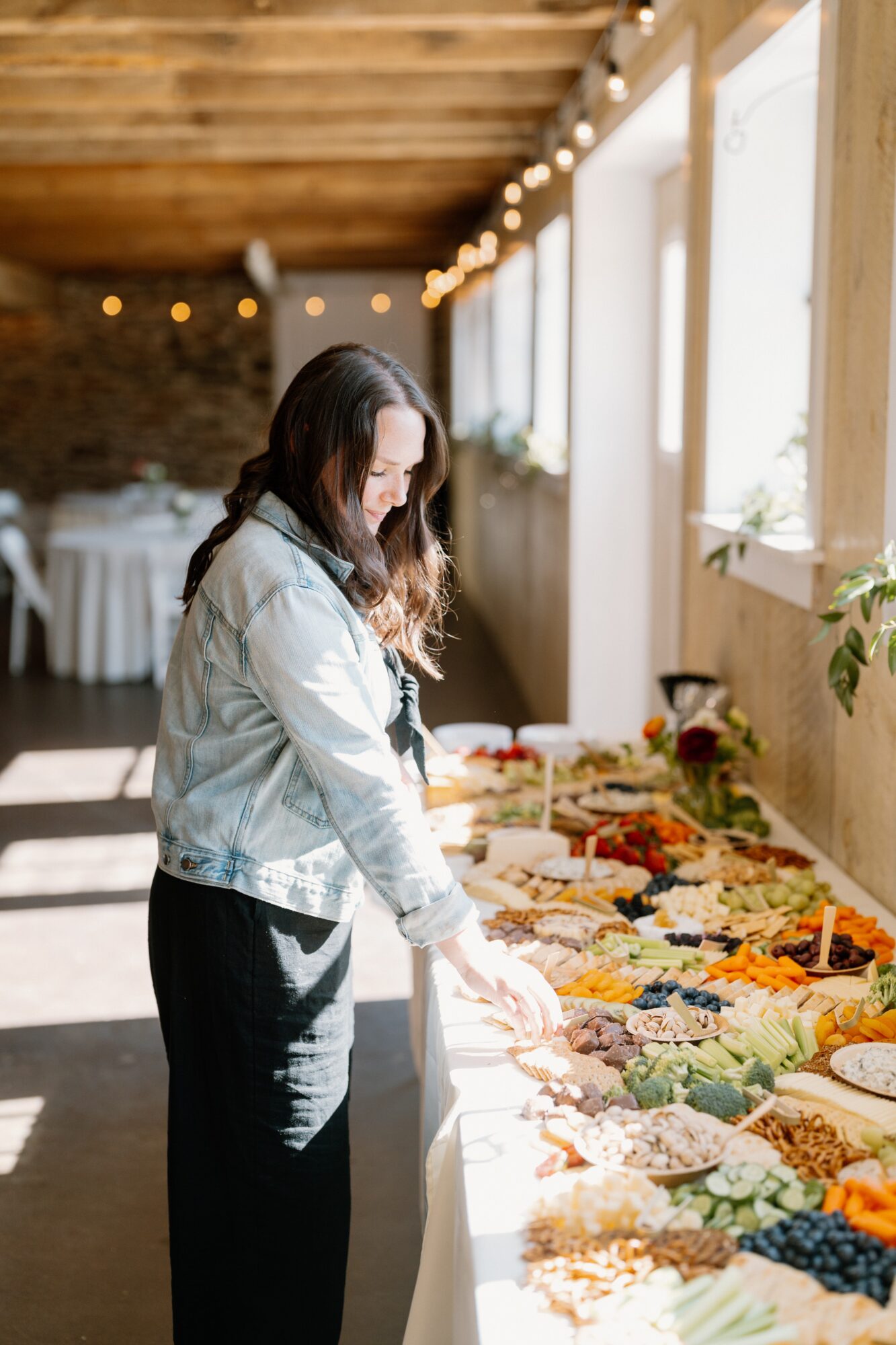 Woman in denim jacket and black pants looking at a table of colorful food in a bright room with large windows.