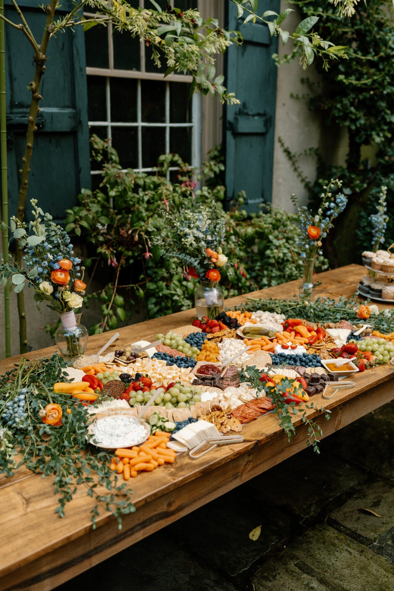 Long outdoor table with food, surrounded by greenery and a window with shutters, decorated with flowers and candles.